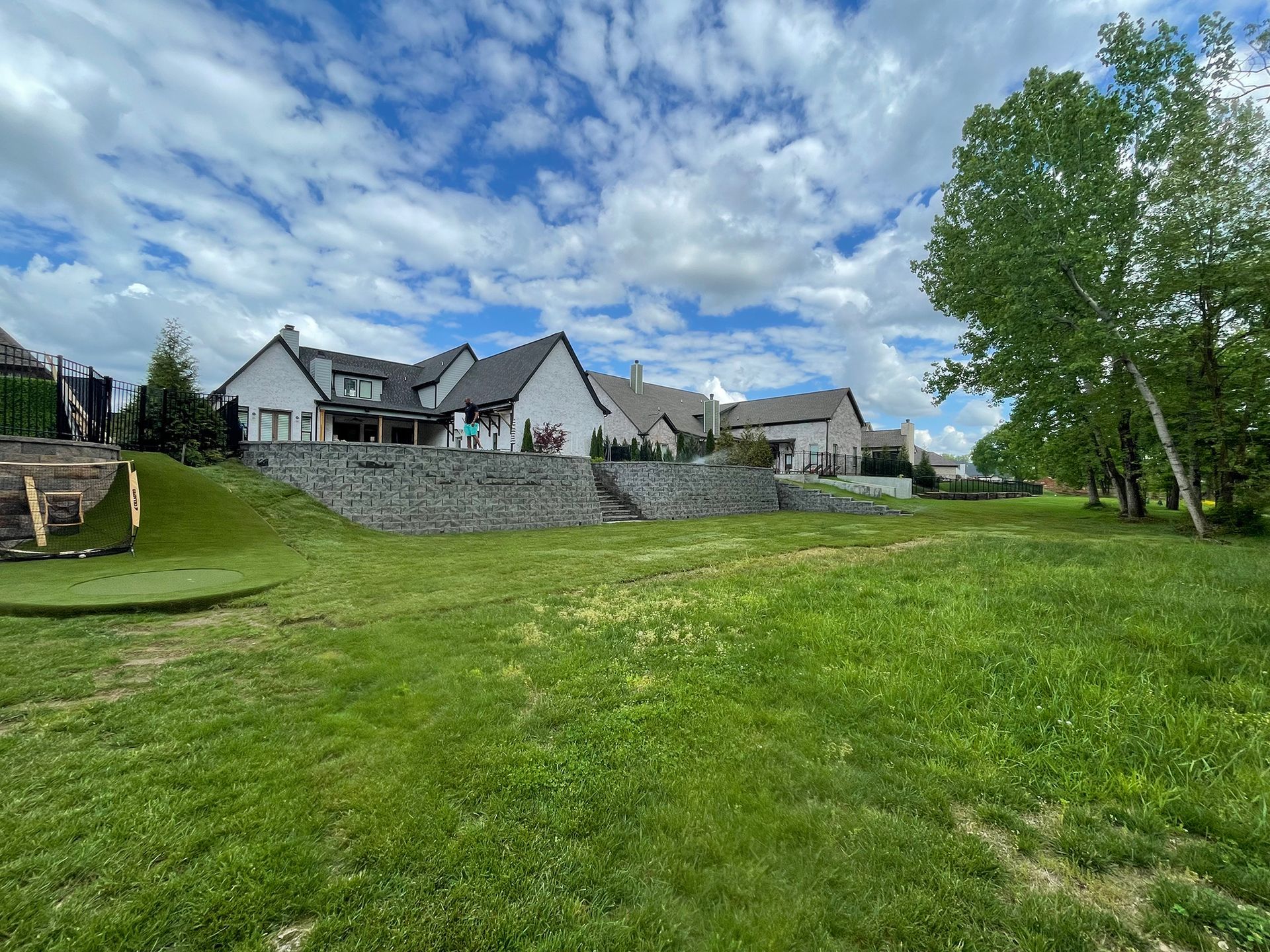 Lush green yard slopes up to a large, modern white house with a retaining wall under a cloudy sky.