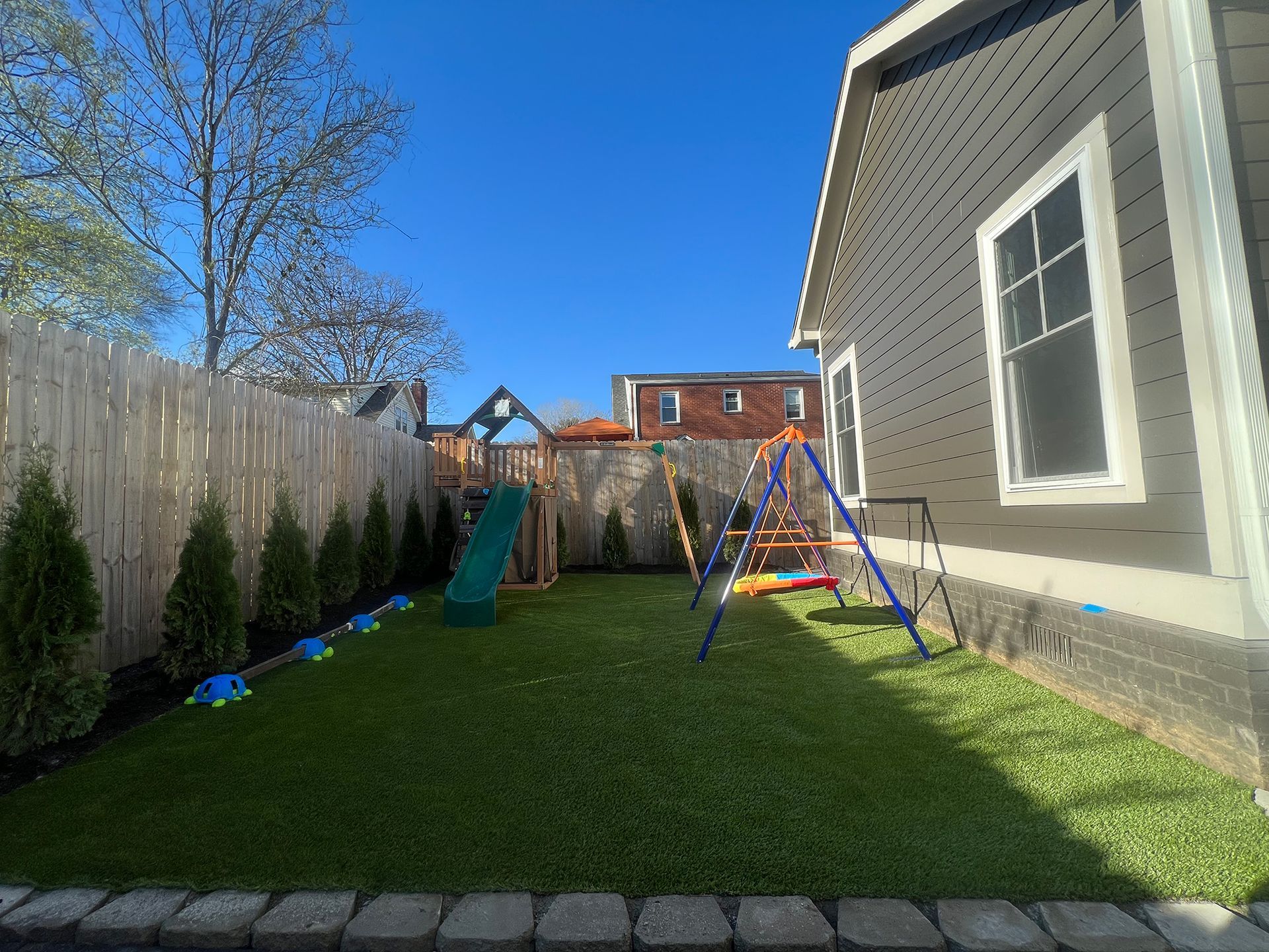 Backyard with a swing set, slide, and play structure, bordered by a fence and house, under a blue sky.