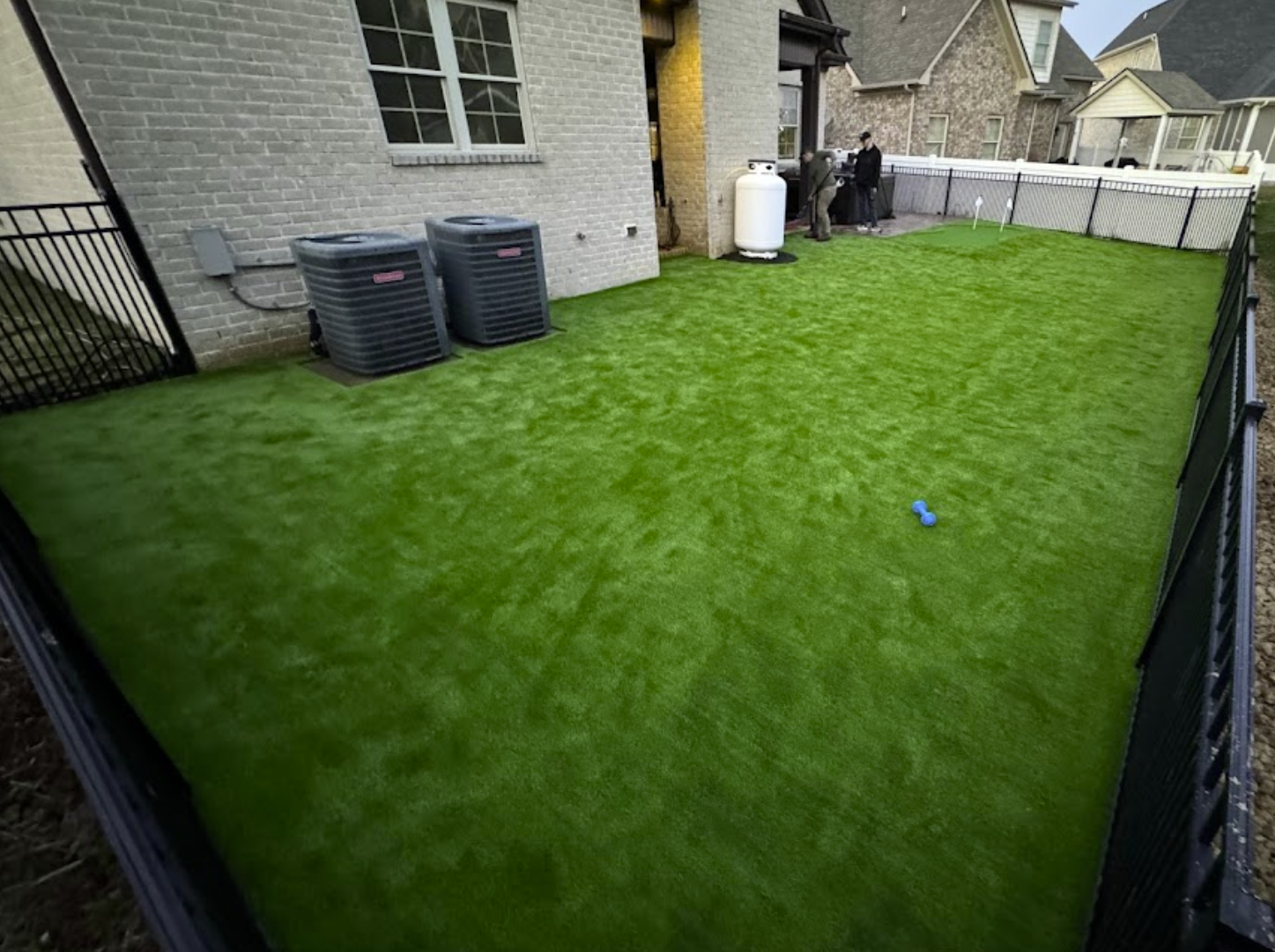 Artificial green lawn in a fenced backyard, next to a brick house and air conditioning units.