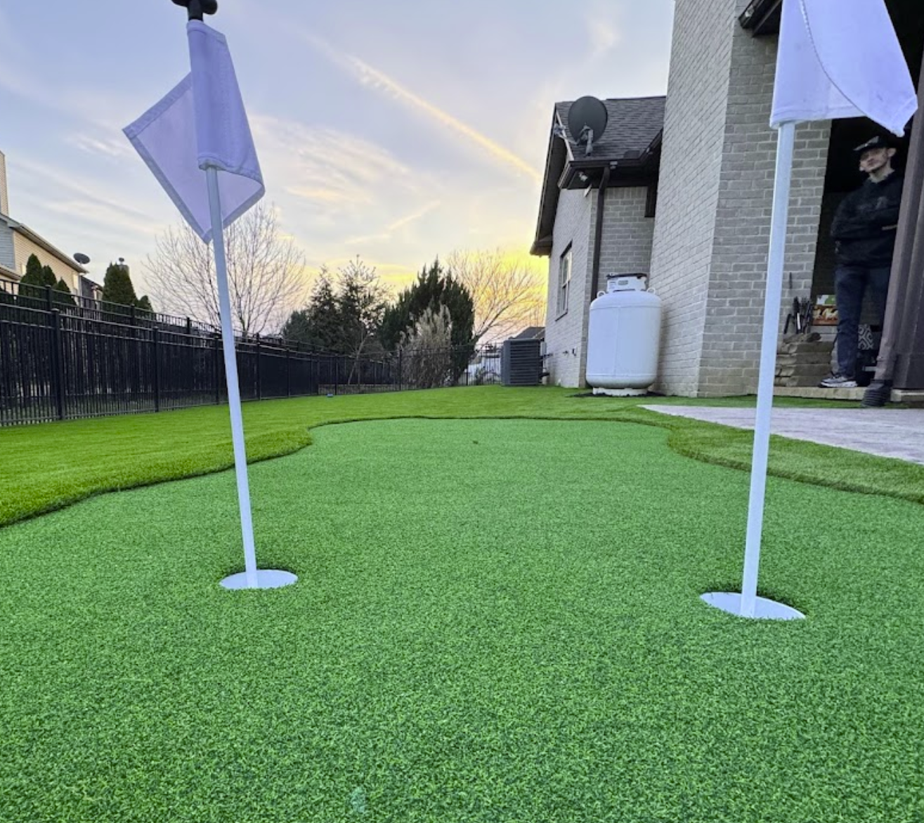 Two white-flagged golf holes on a backyard putting green; a person stands nearby.