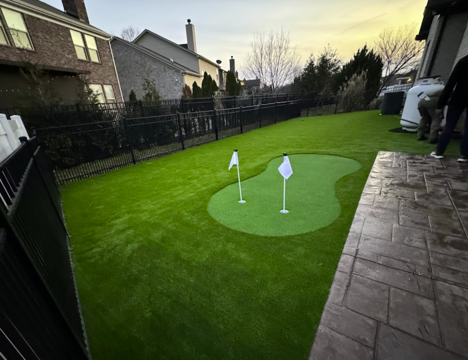 Backyard putting green with two flags on green turf, surrounded by fence and houses.