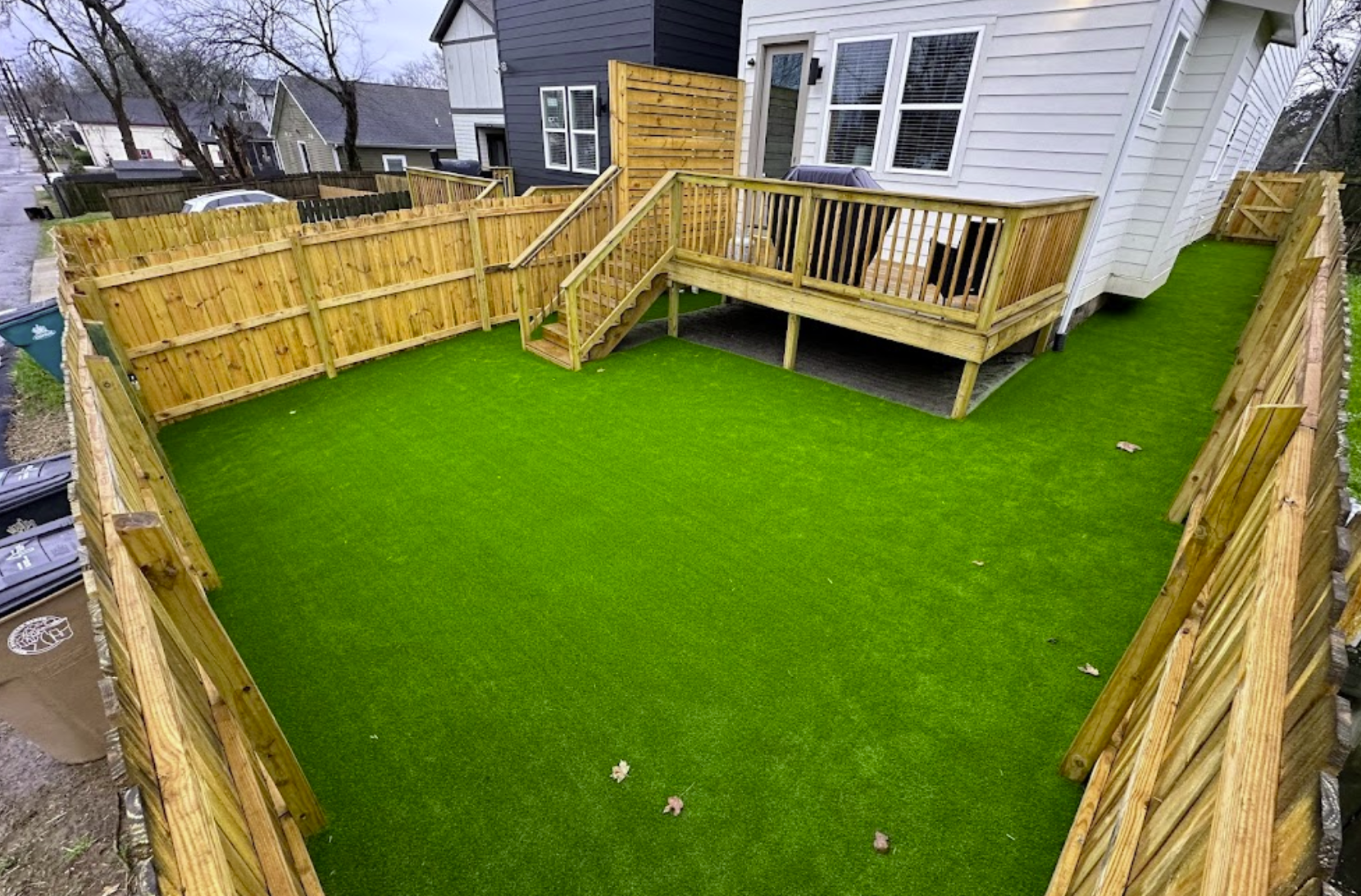 A fenced-in backyard with bright green artificial grass and wooden deck connected to a white house.