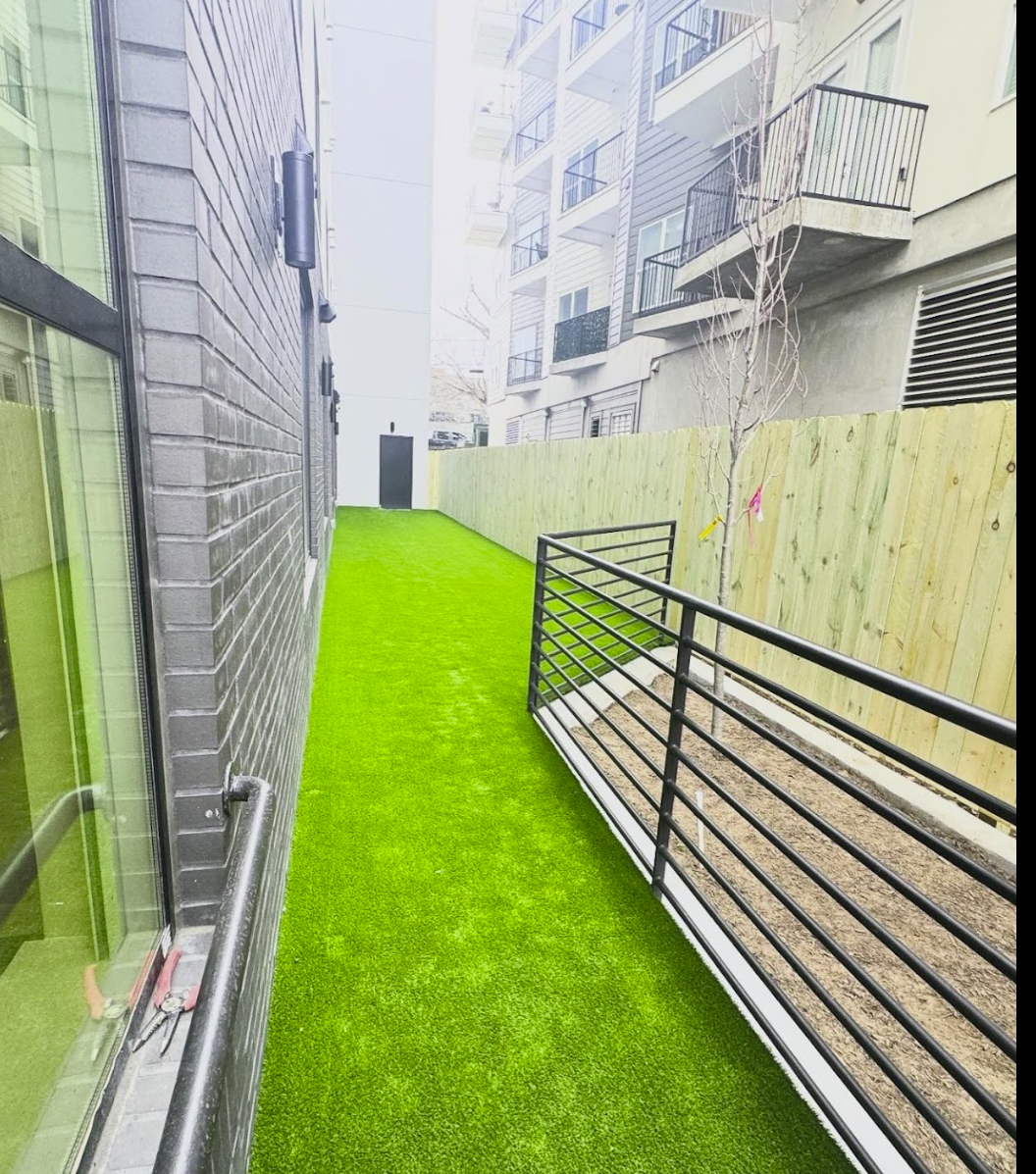 A narrow alley with artificial green turf between a brick building and a wooden fence.
