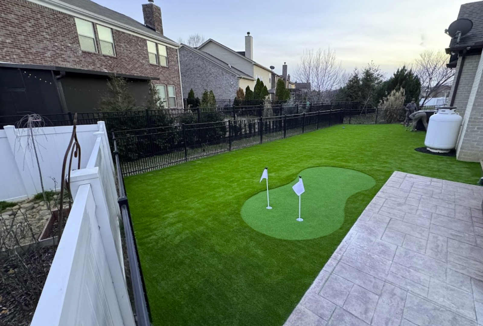 Backyard with golf putting green, surrounded by black fence, white fence, and two-story houses.