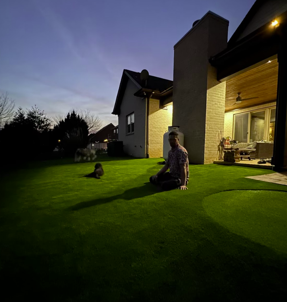 Man and small dog sit on green lawn at dusk, near a house with lights on.