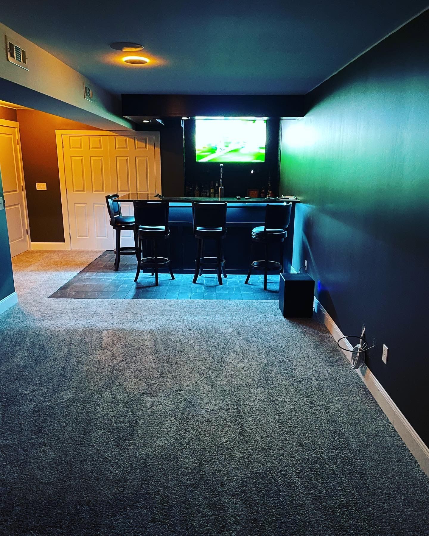 Basement bar with stools, TV, and dark blue walls. Carpet floor and white doors are visible.