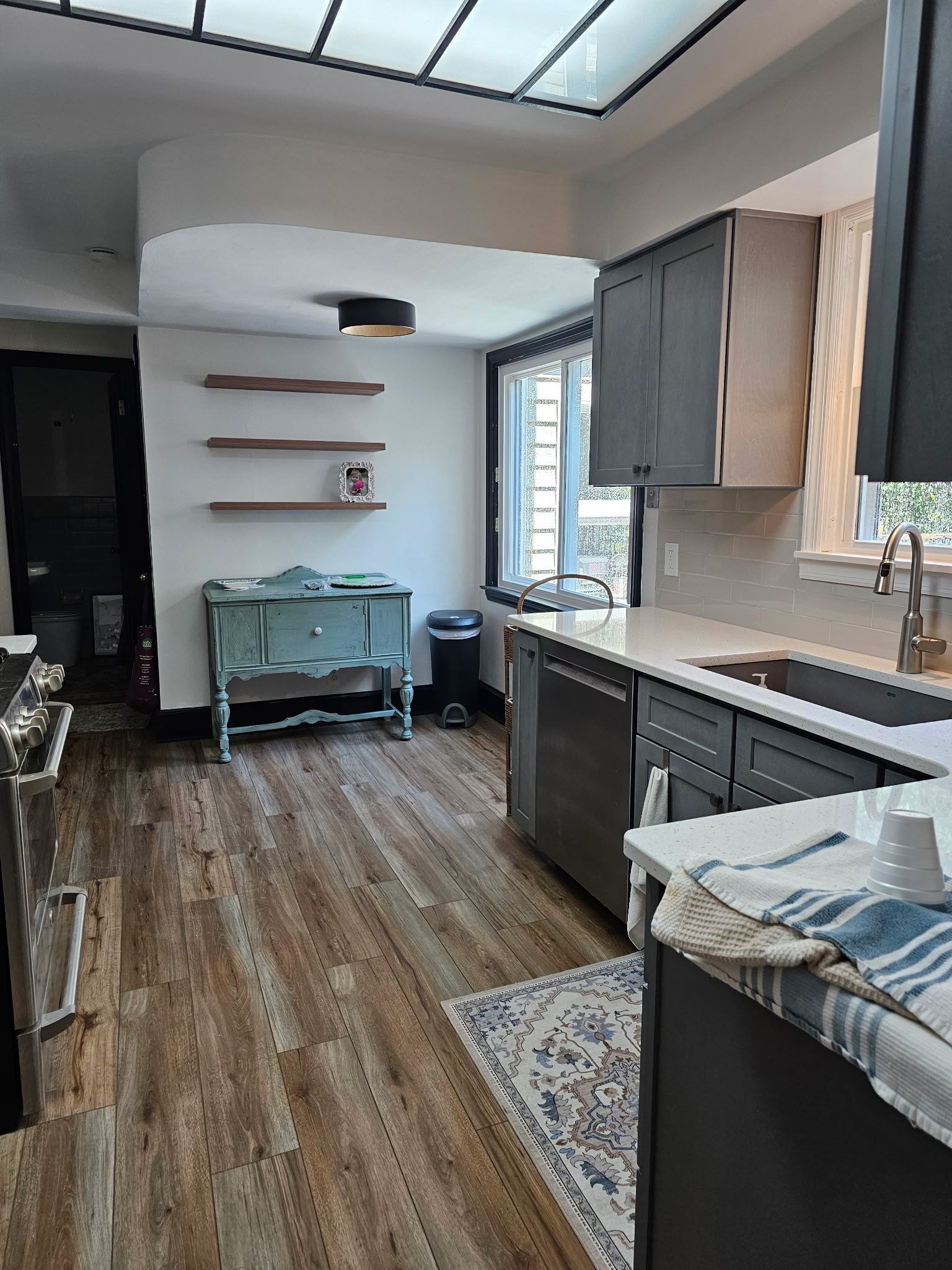 Kitchen with gray cabinets, wooden floor, and a light-filled skylight. Includes a teal cabinet and floating shelves.
