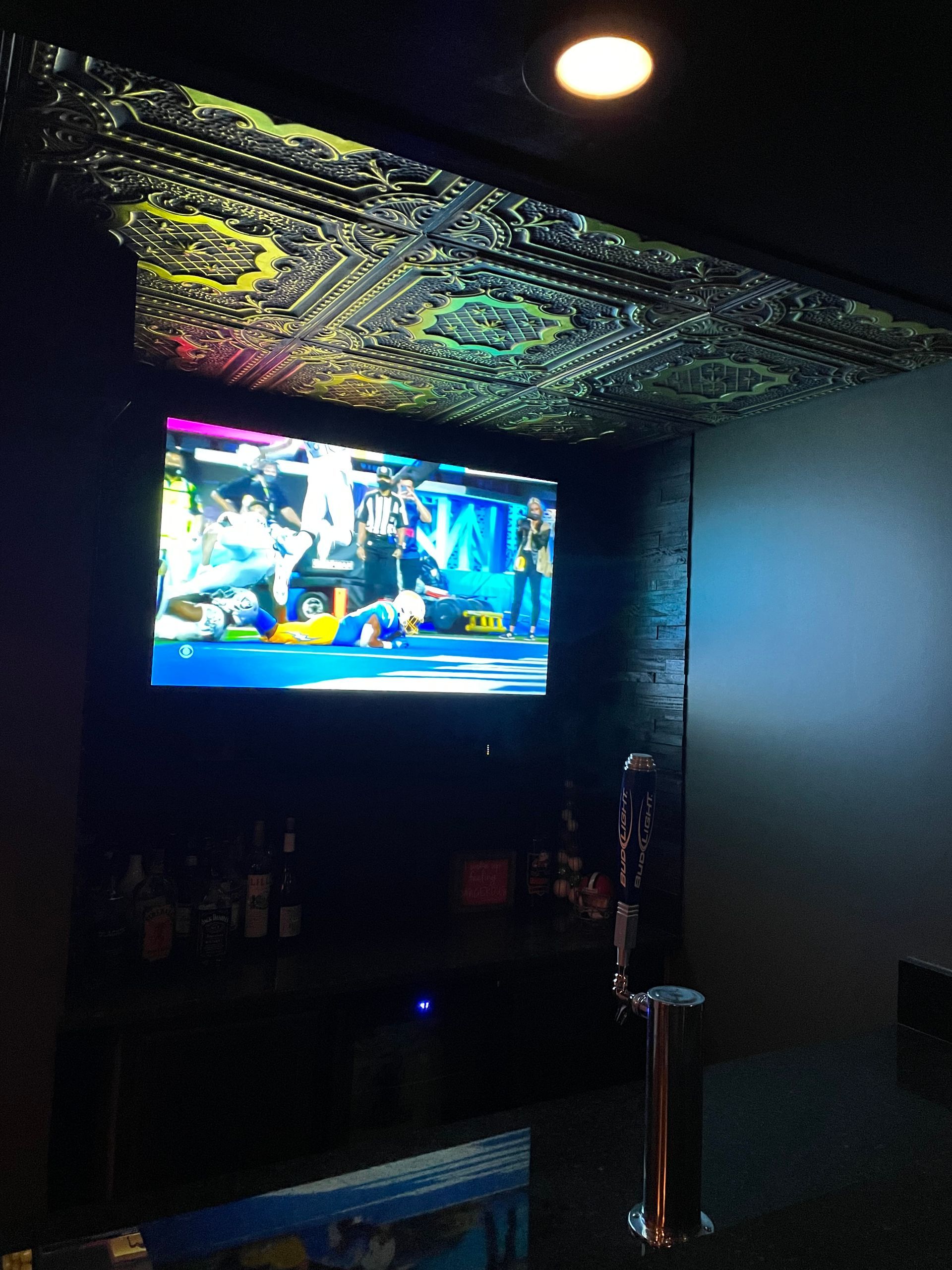 TV screen displaying a video game above a bar; decorative ceiling, dimly lit.