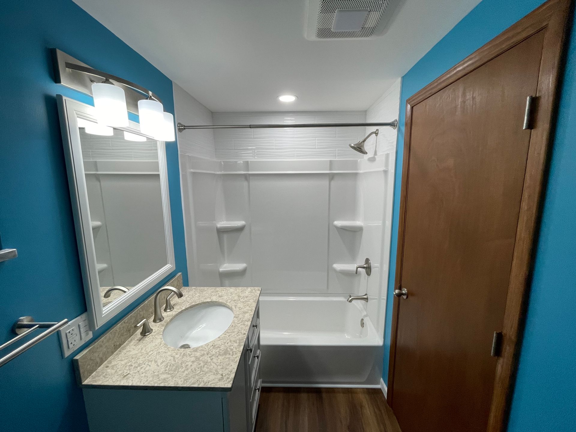 Bathroom with blue walls, white tub and shower, vanity with light-colored countertop and sink, and brown door.