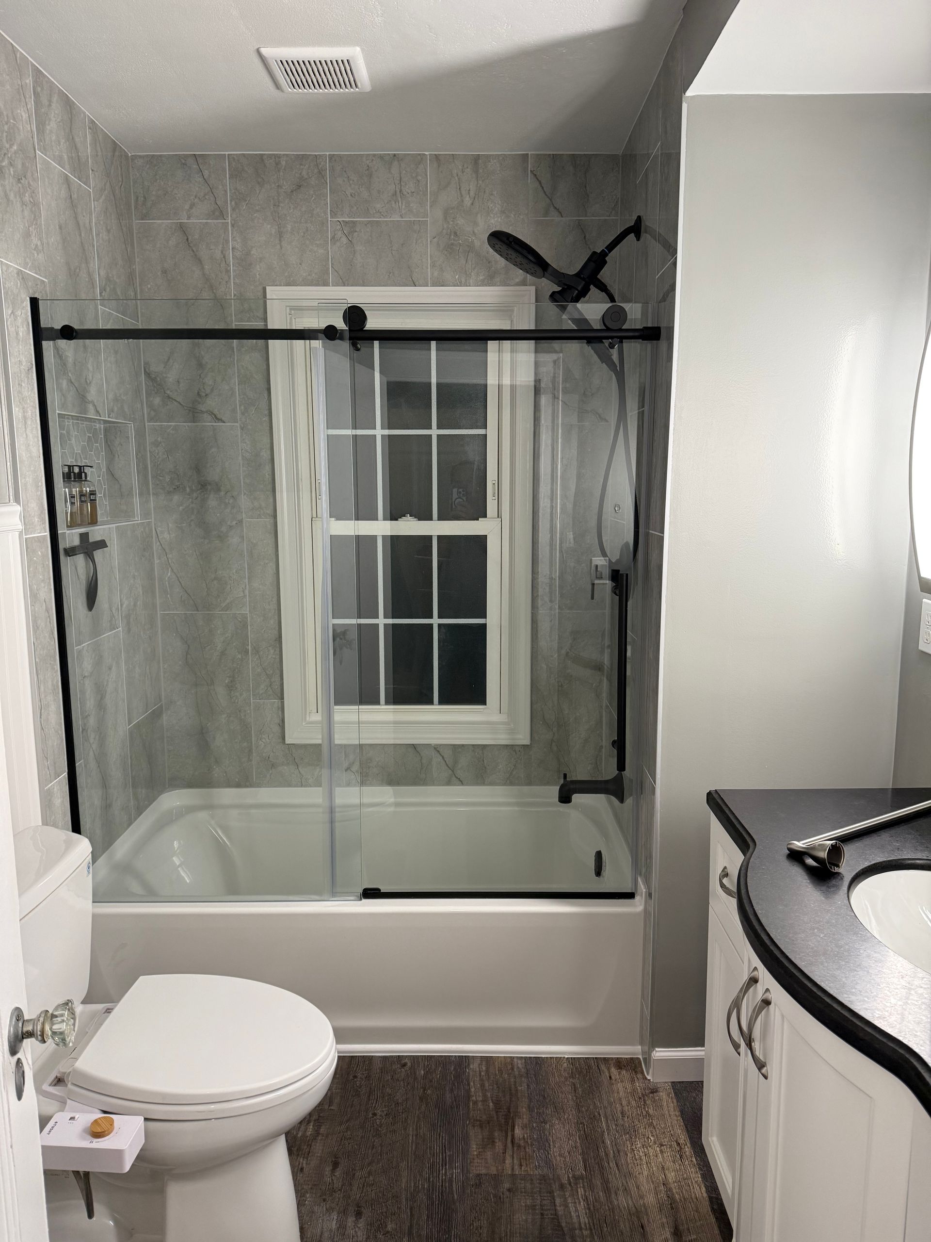 Bathroom with gray tile, a tub, glass shower door, and a toilet.