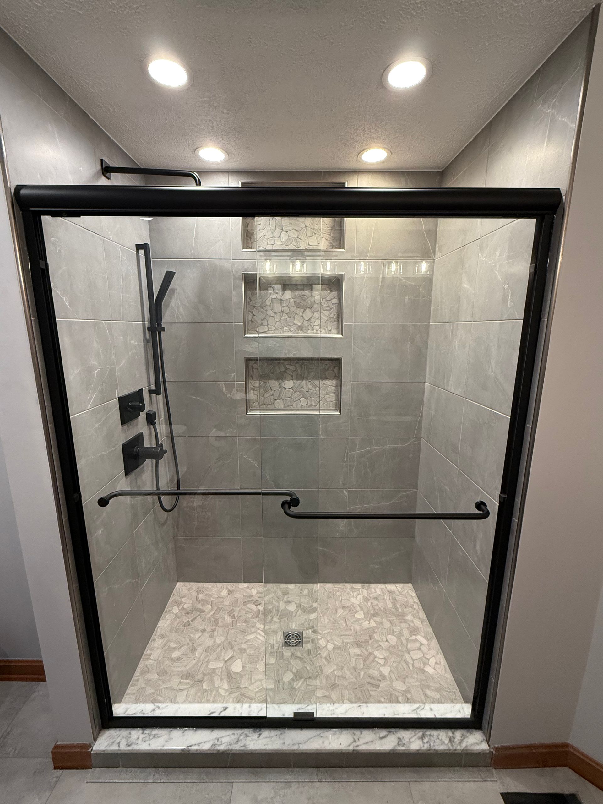 Gray tiled shower with glass door and black fixtures. Built-in shelves.