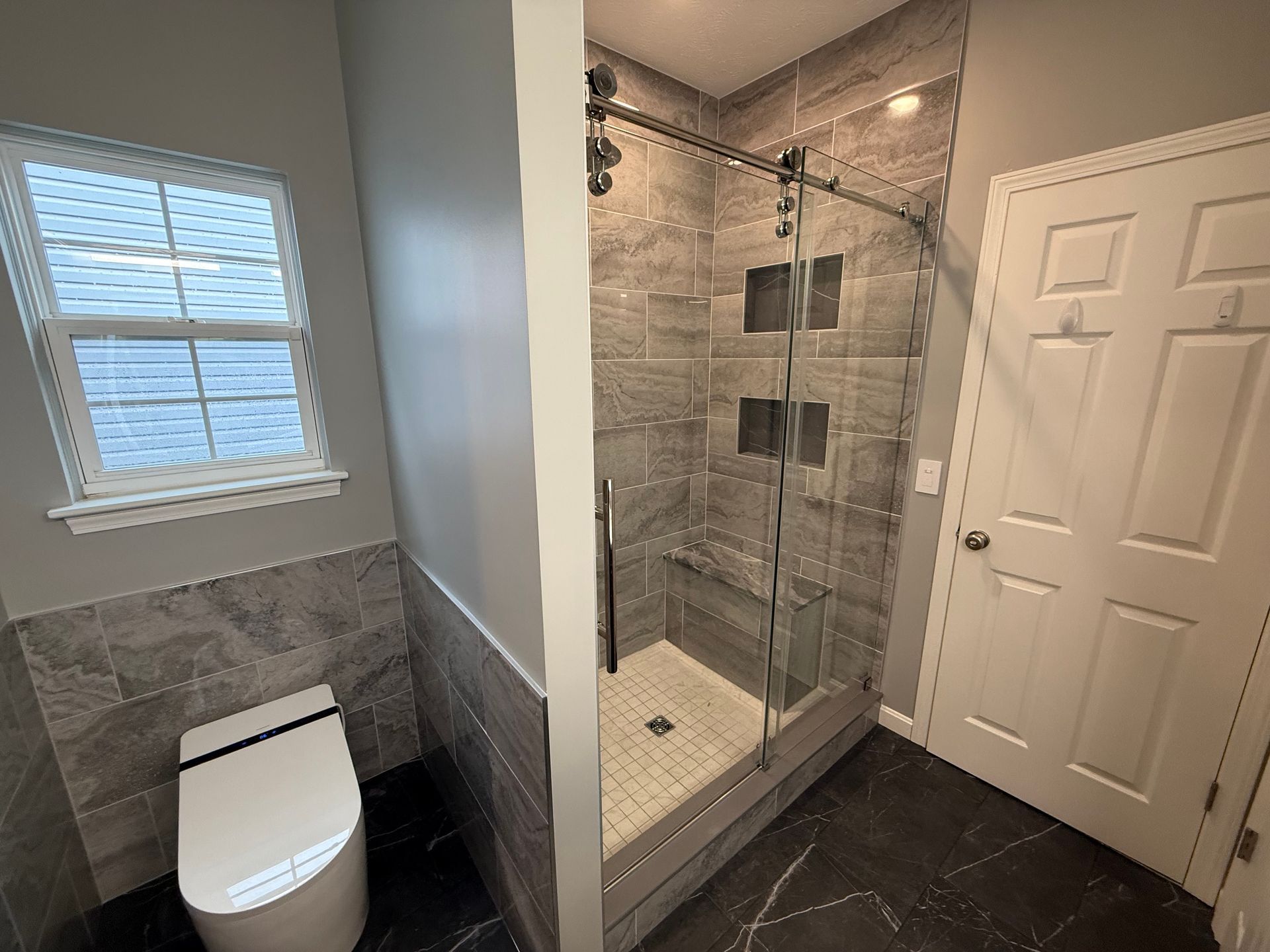Bathroom with tiled walls, glass shower, toilet, and a window.