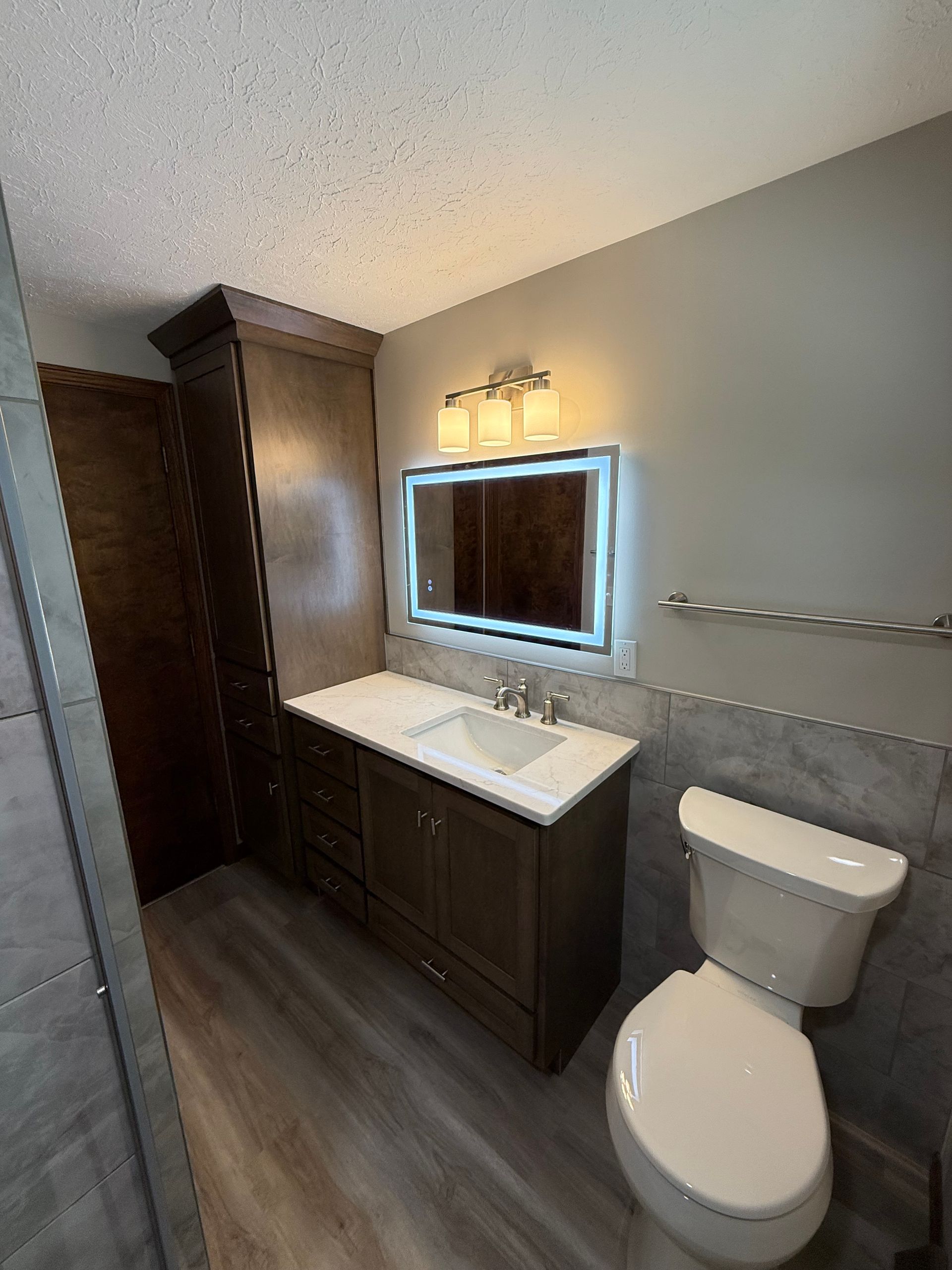 Bathroom with dark brown vanity, lit mirror, tall cabinet, and gray walls.
