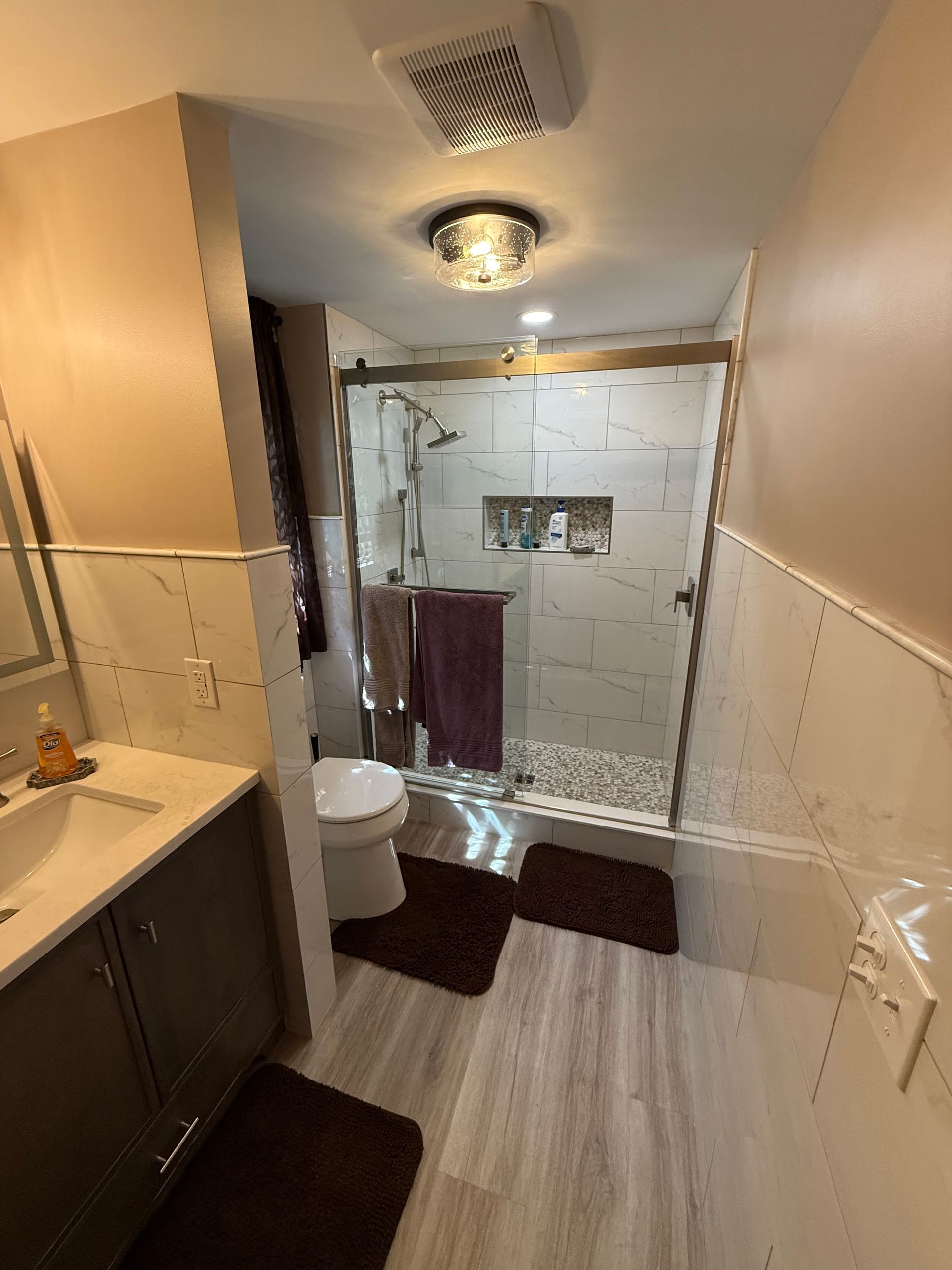 Small bathroom with sink, toilet, and glass shower enclosure; beige walls and dark floor mats.