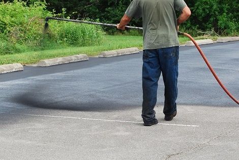 A person wearing a grey shirt and blue jeans sprays asphalt sealer onto a parking lot surface.