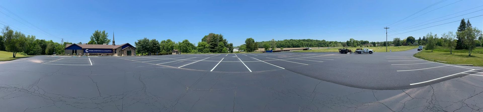 A wide-angle shot of a nearly empty, paved parking lot under a clear blue sky, with trees and a building in the distance.