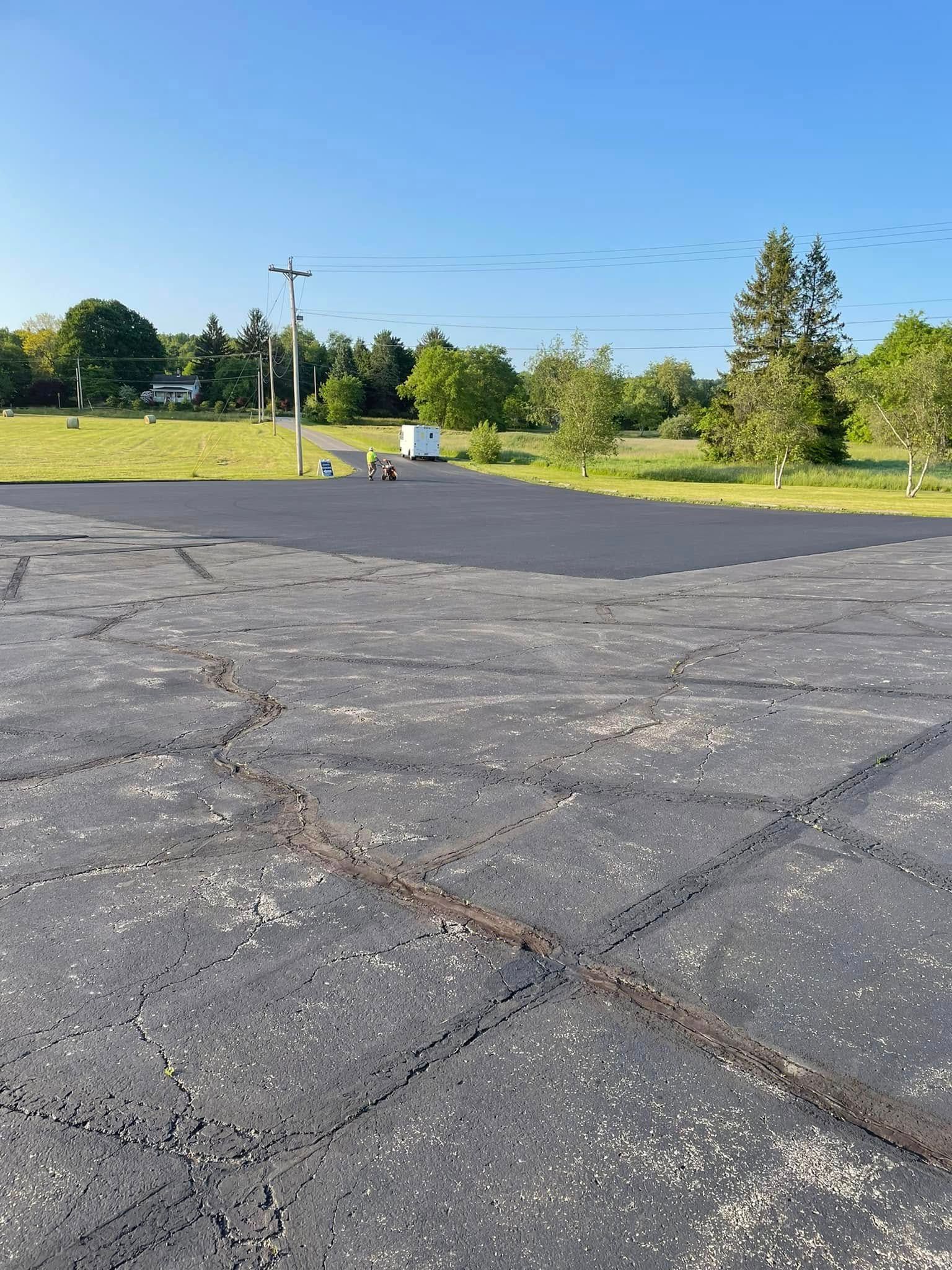 An asphalt parking lot showing a contrast between older, cracked pavement in the foreground and a newly paved section.