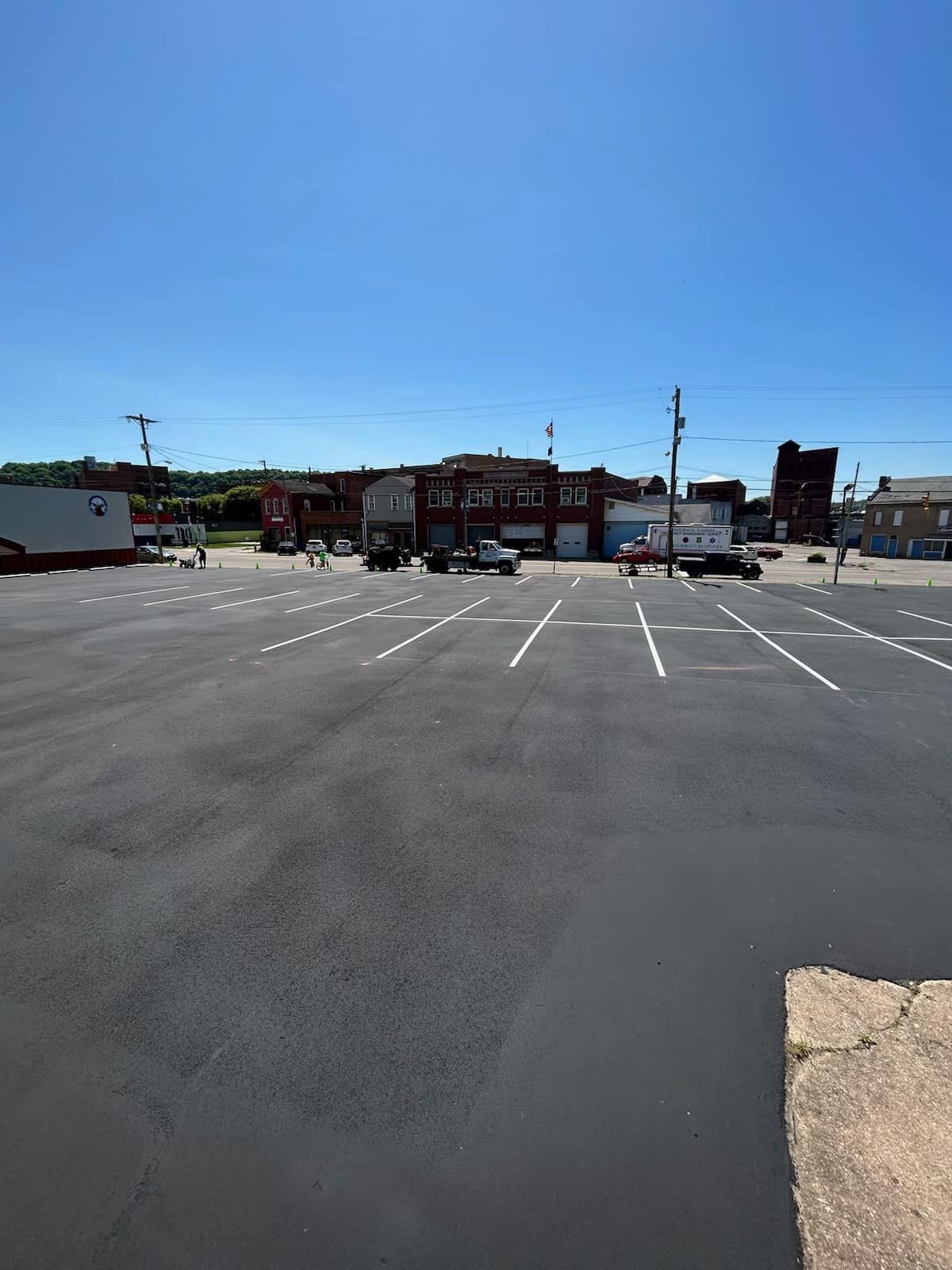 A large, empty parking lot under a bright blue sky, with older brick buildings and utility poles in the background.