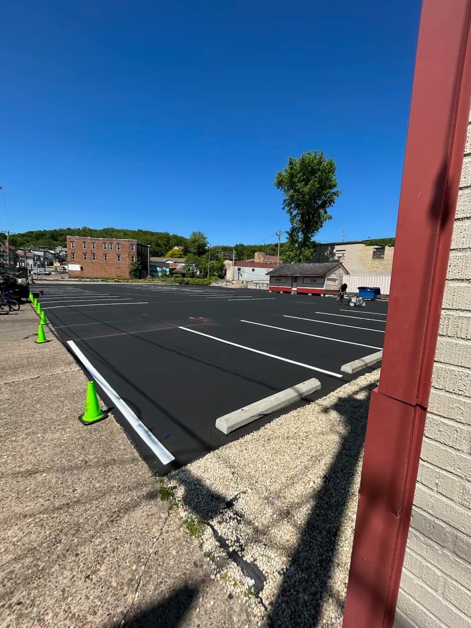 A newly paved parking lot with white-painted lines and concrete parking stops, bordered by gravel and a red building corner.