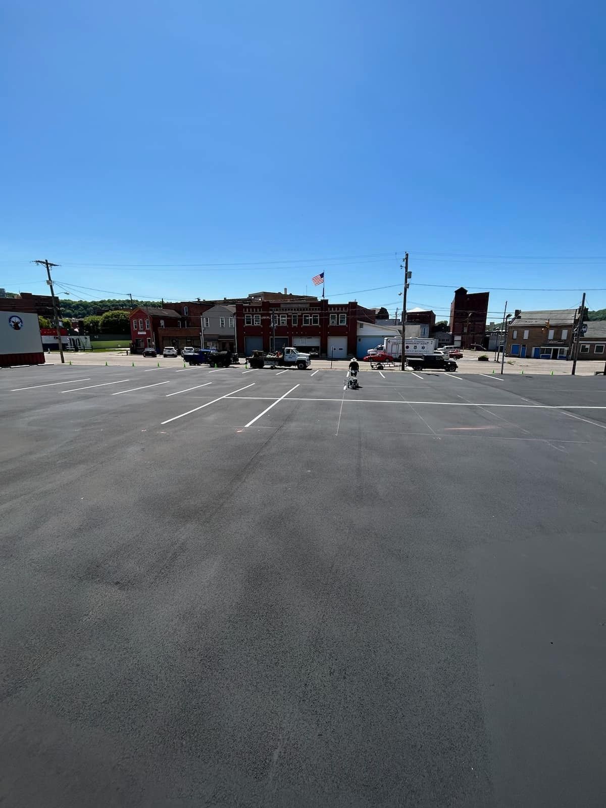 A wide, empty asphalt parking lot under a clear blue sky, with commercial buildings and parked vehicles in the distance.