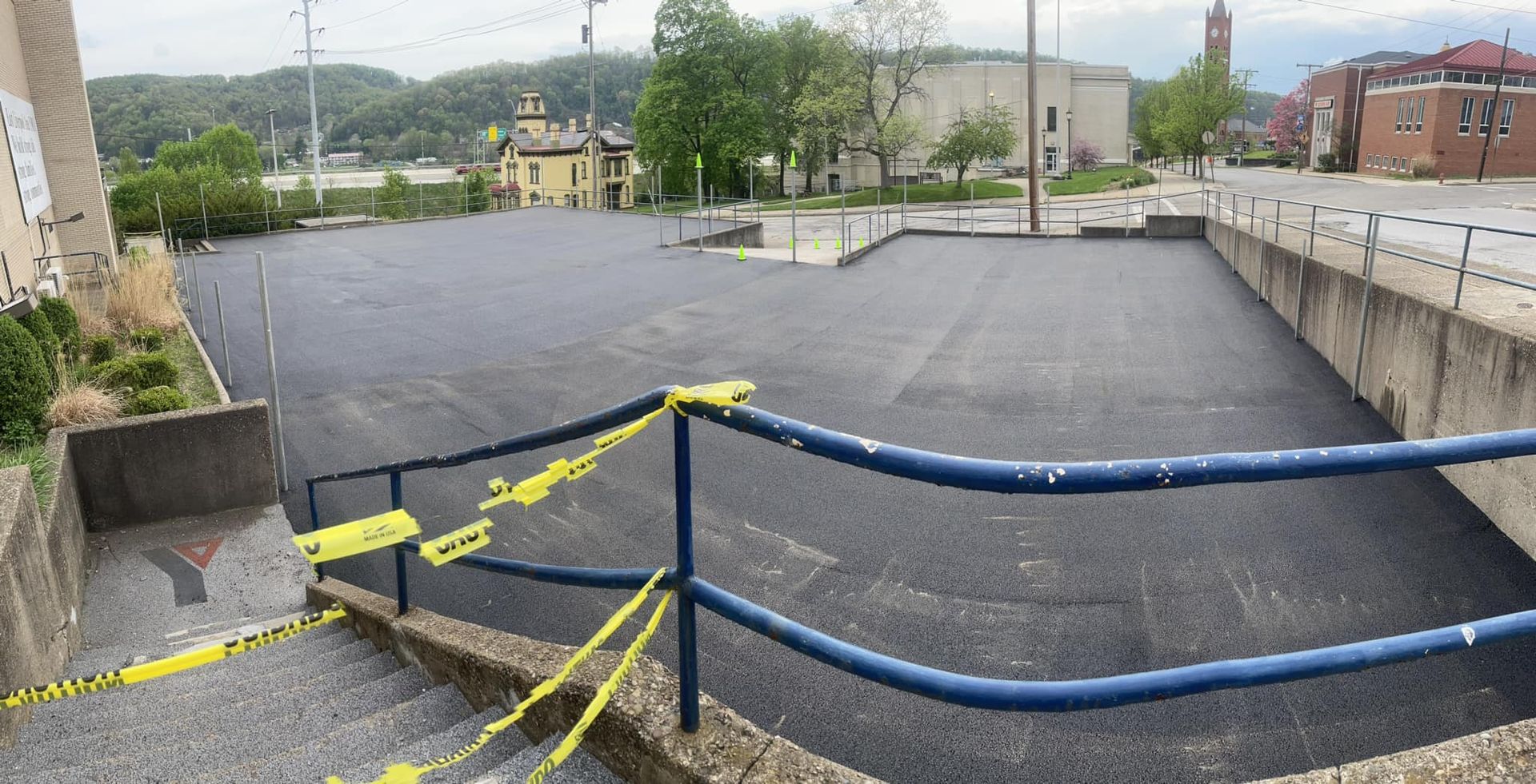 Newly paved black asphalt parking lot with blue metal railings and yellow caution tape at the top of concrete stairs.