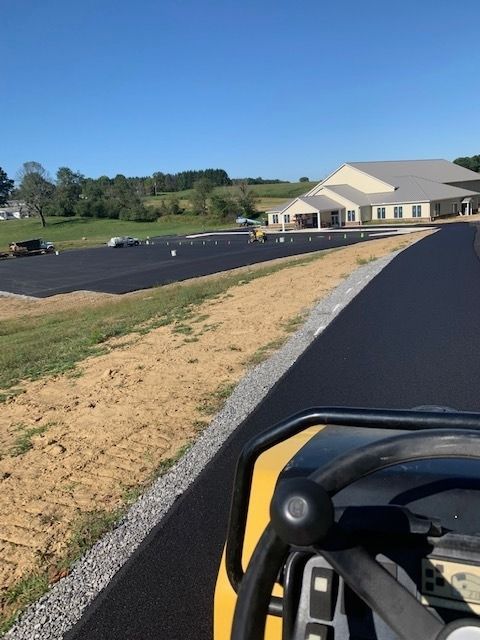 View from a paving vehicle showing a freshly paved black asphalt parking lot leading toward a large, light-colored building.