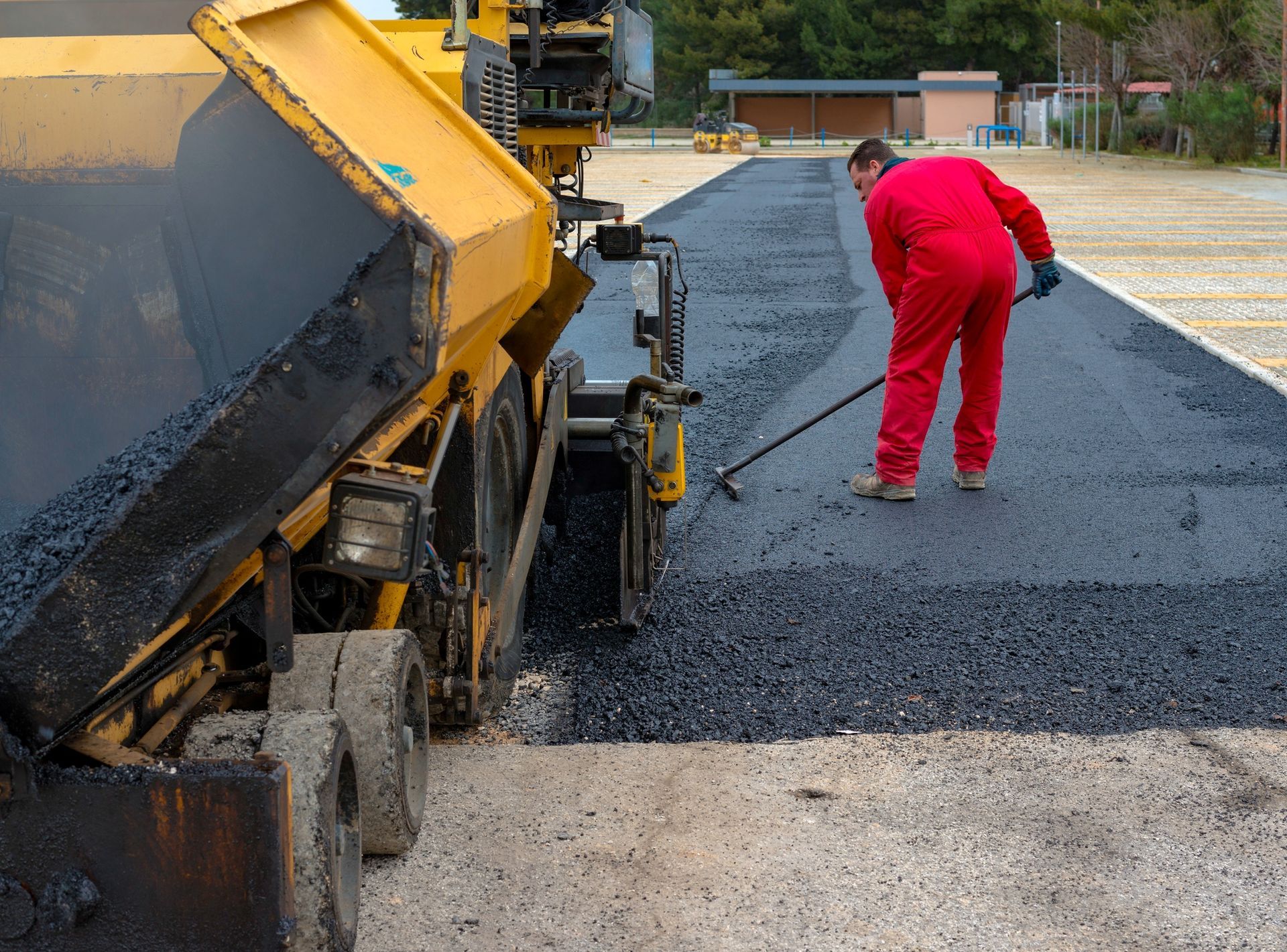 A worker in a red jumpsuit uses a hand tool to spread fresh black asphalt laid by a yellow paving machine.