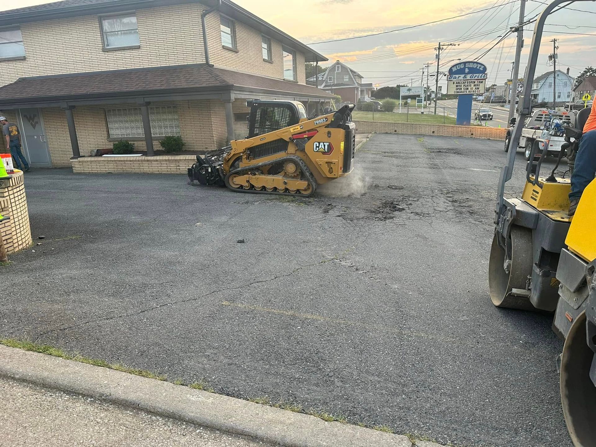 A yellow tracked skid steer clears gravel in a parking lot next to a tan building at dusk.