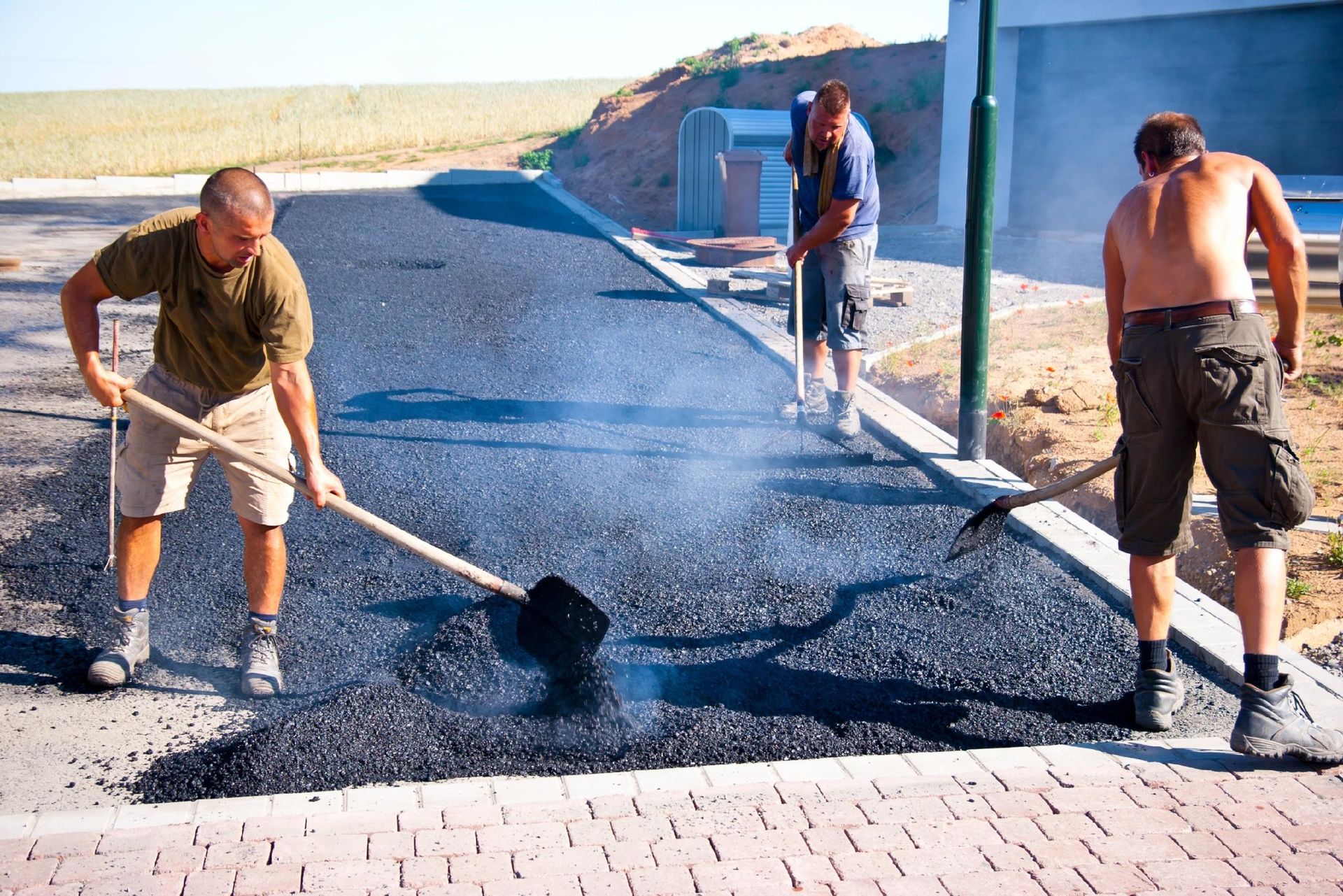 Three workers use shovels to spread hot asphalt on a driveway, with steam rising from the material.