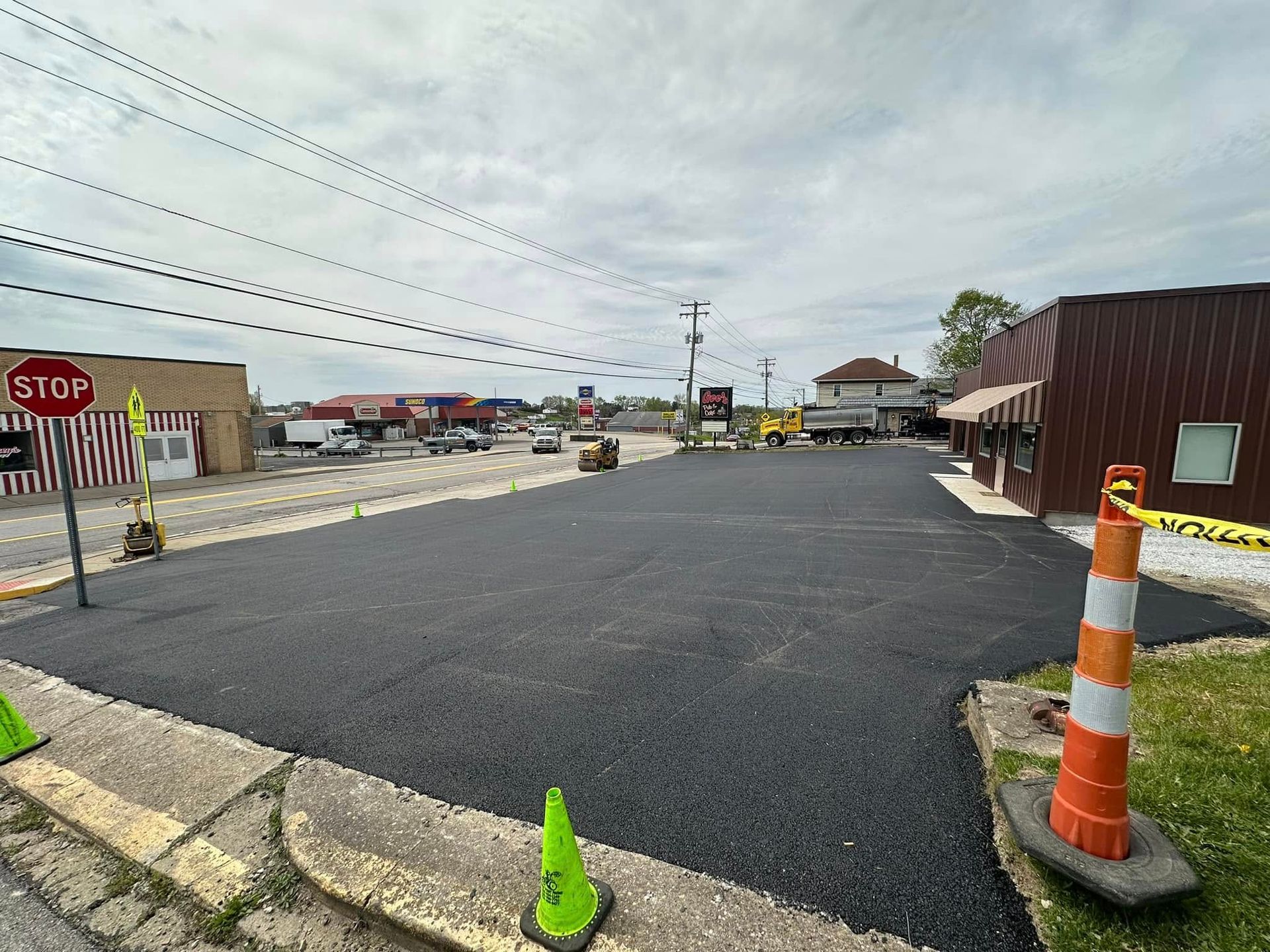A freshly paved asphalt parking lot on a cloudy day, bordered by a sidewalk, orange traffic cones, and a nearby building.