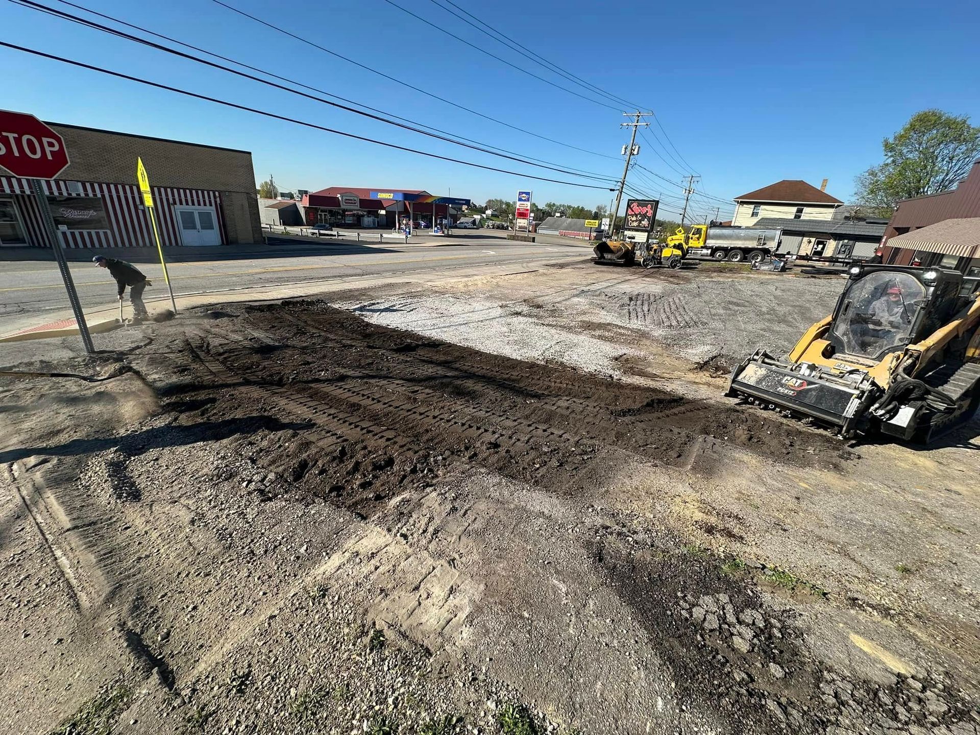 A yellow skid steer loader sits on a gravel construction site under a clear blue sky, with a nearby building and stop sign.