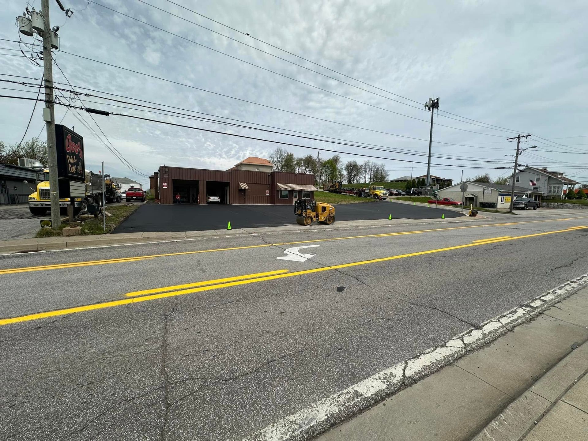 A yellow steamroller compacts fresh black asphalt in front of a brick commercial building on a street.