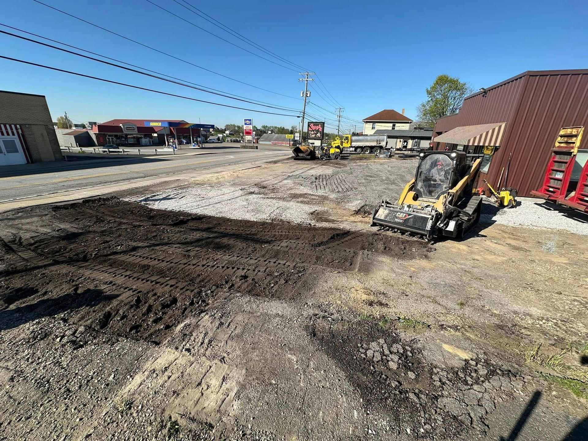 Construction site with a yellow skid steer loader on gravel and dirt, adjacent to a brown building under a clear blue sky.