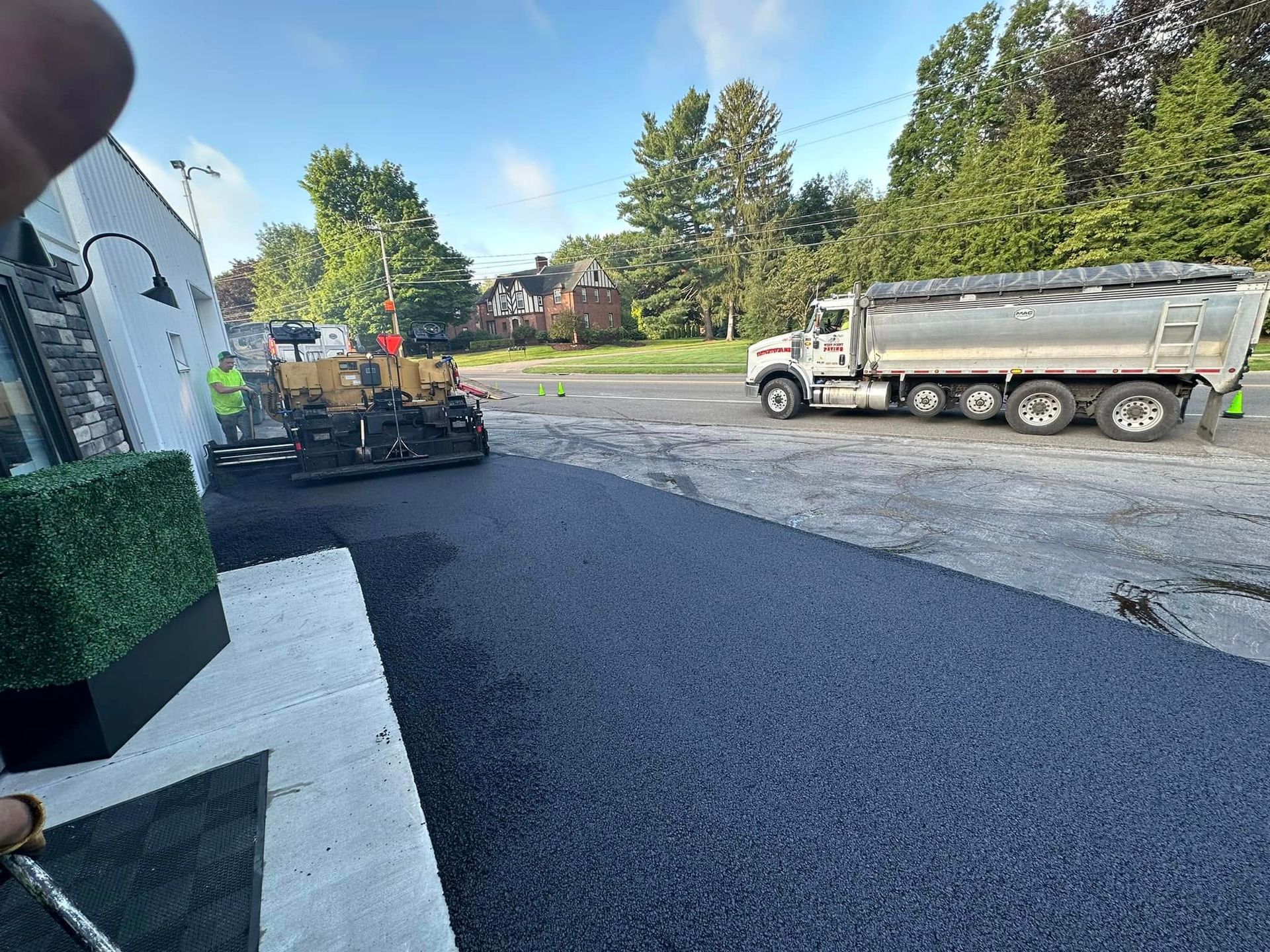 Construction crew paving a dark asphalt driveway next to a white building, with a dump truck parked nearby.