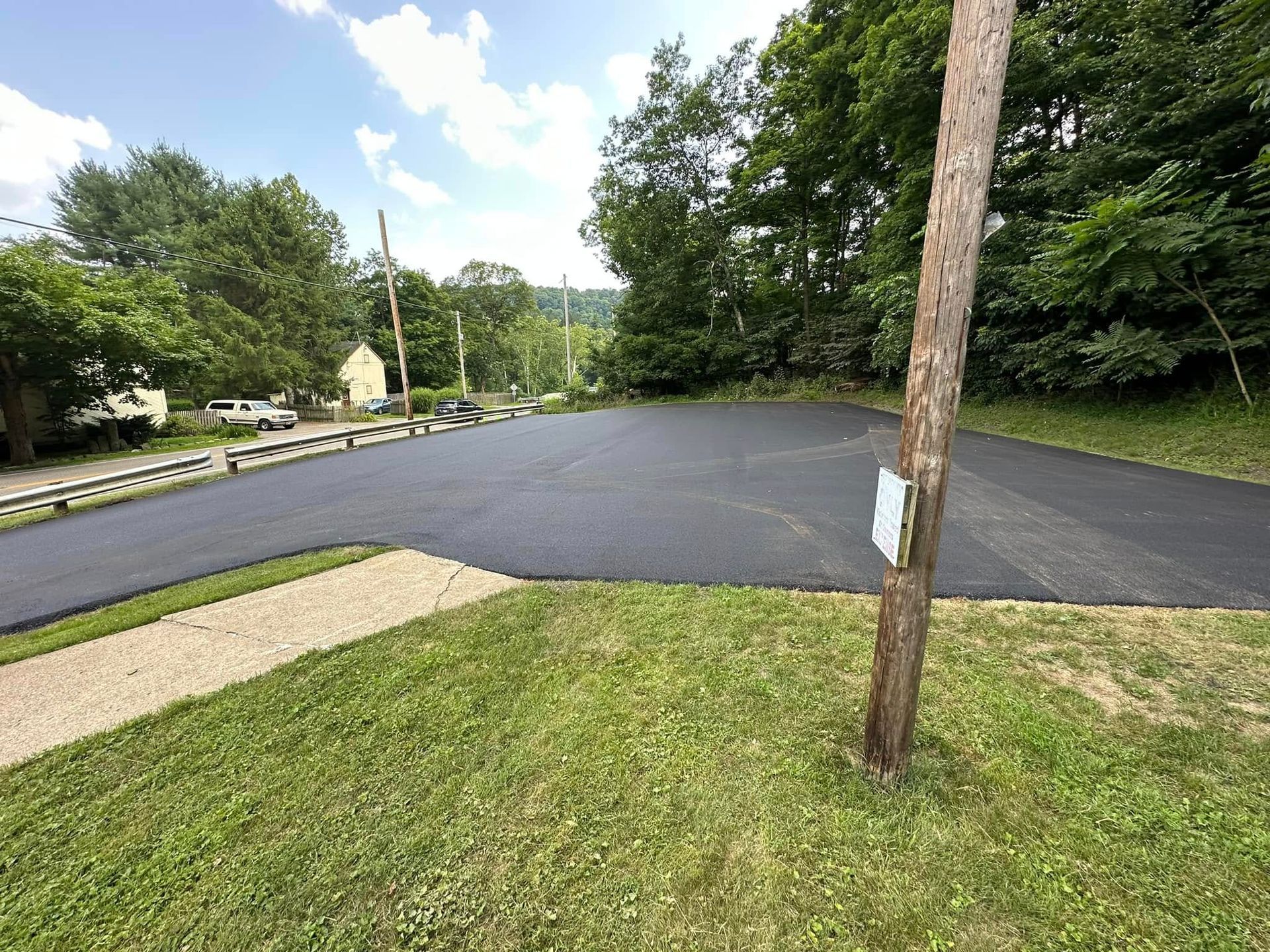 A freshly paved asphalt parking lot sits next to a grassy lawn under a sunny, partly cloudy sky.