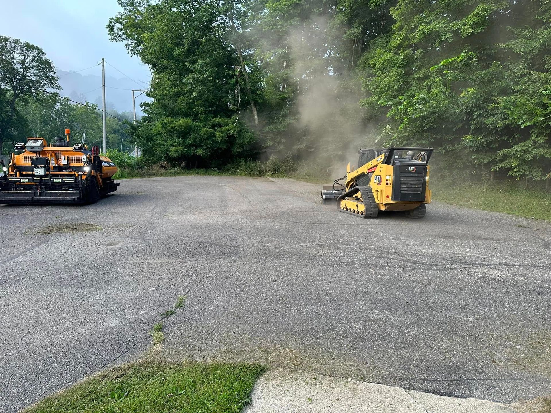 A yellow skid steer loader creates a cloud of dust while working on a gravel lot near an asphalt paving machine.