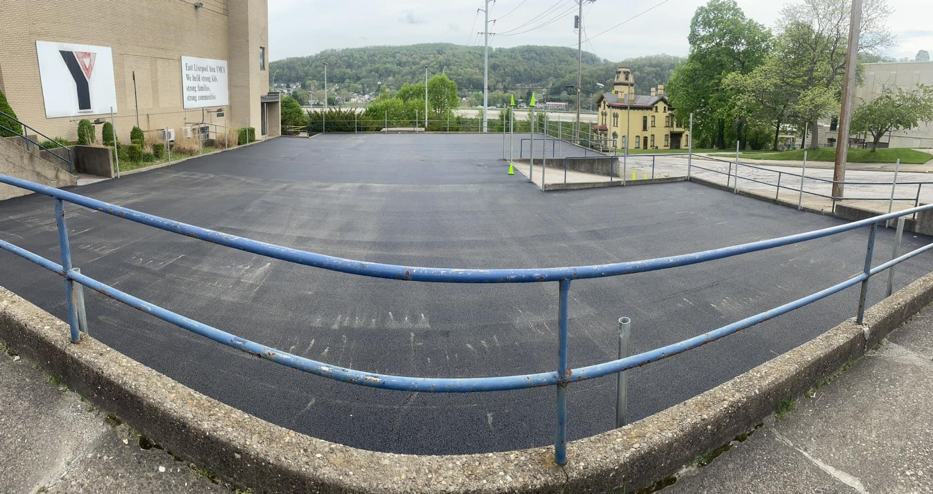 A freshly paved, empty black asphalt parking lot framed by a blue metal railing in front of a YMCA building.