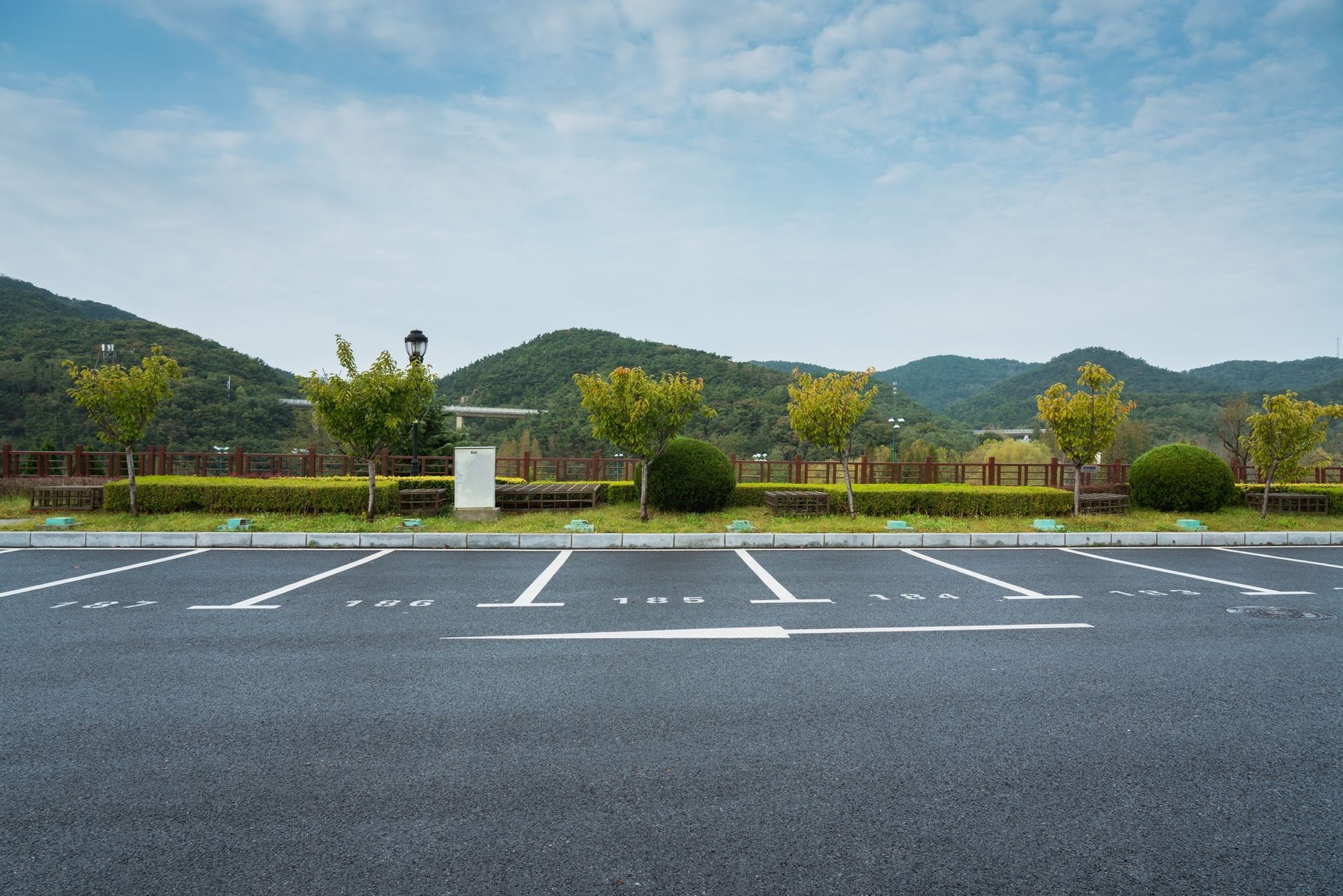 An empty asphalt parking lot with painted lines, framed by a manicured hedge, trees, and rolling hills under a blue sky.