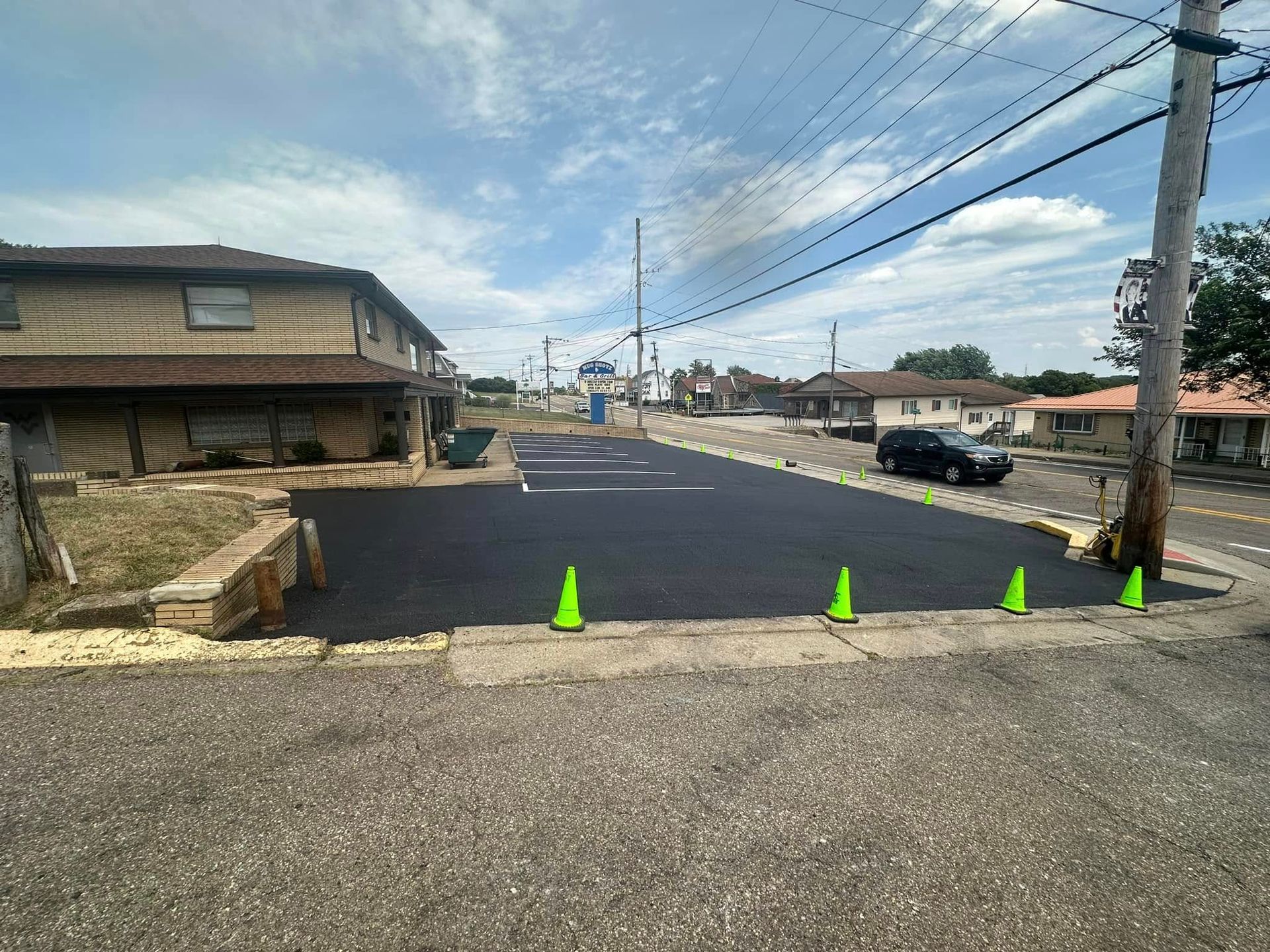 A freshly paved asphalt parking lot with bright green traffic cones lining the edge near a house and a utility pole.