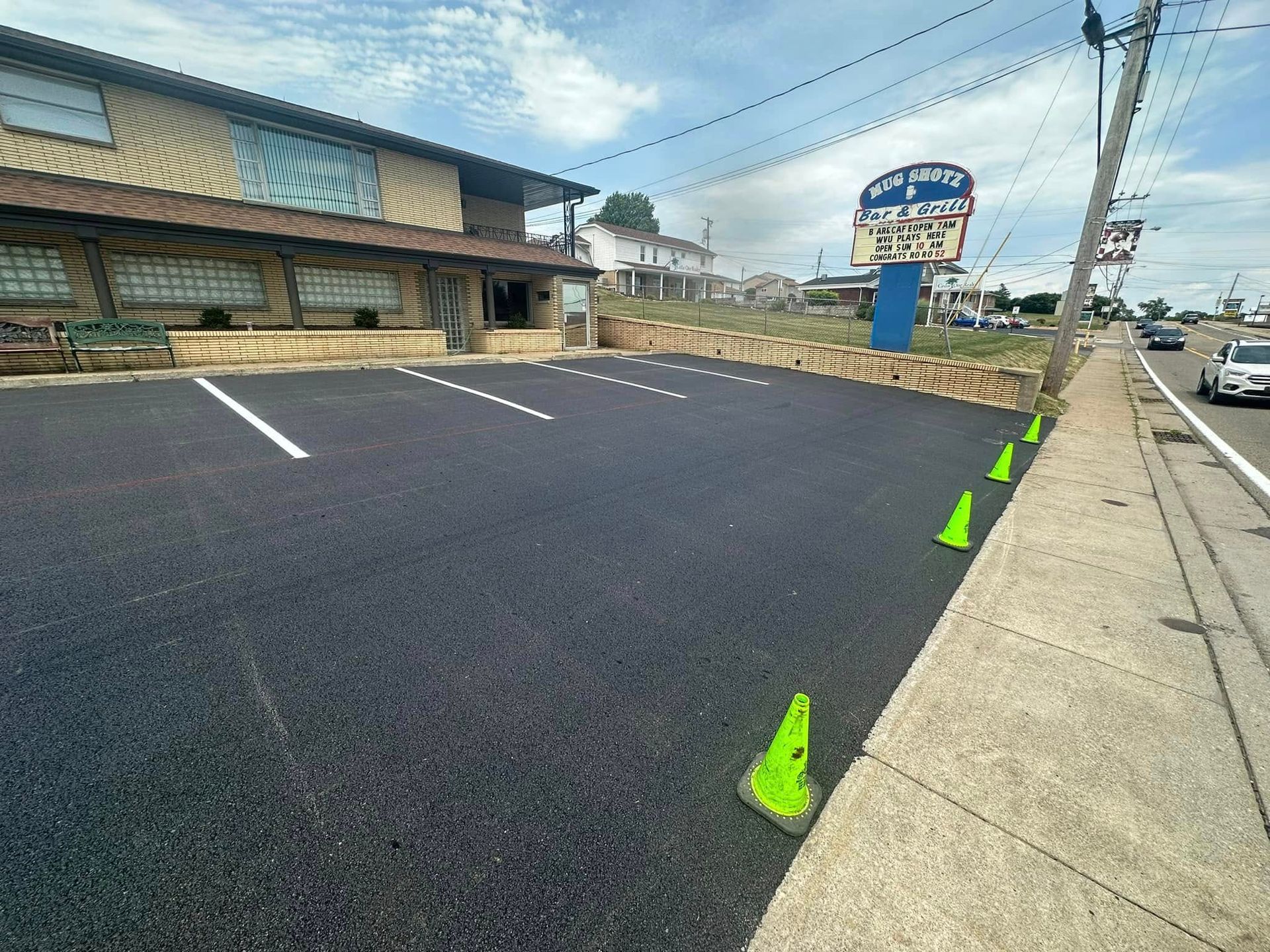 Newly paved parking lot with white painted lines and neon green traffic cones beside a building under a bright sky.