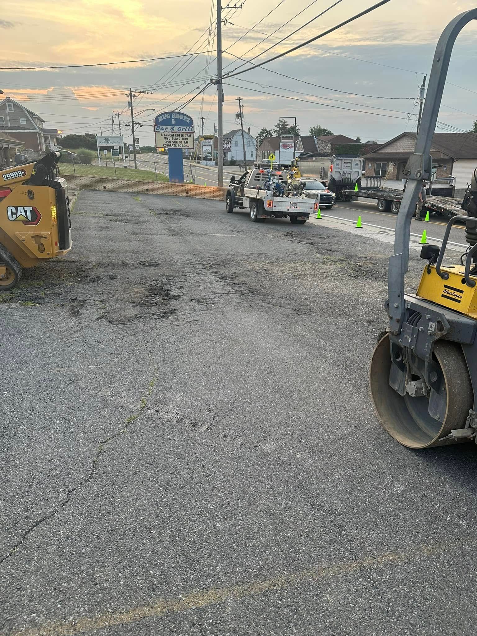 Construction site with heavy machinery, including a yellow CAT skid steer and a steamroller, on a gravel surface.