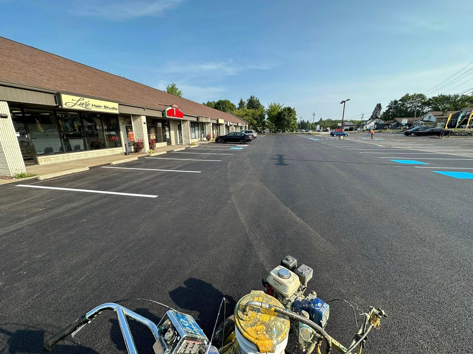 A line-striping machine sits on a freshly paved parking lot in front of a commercial shopping plaza under a clear sky.