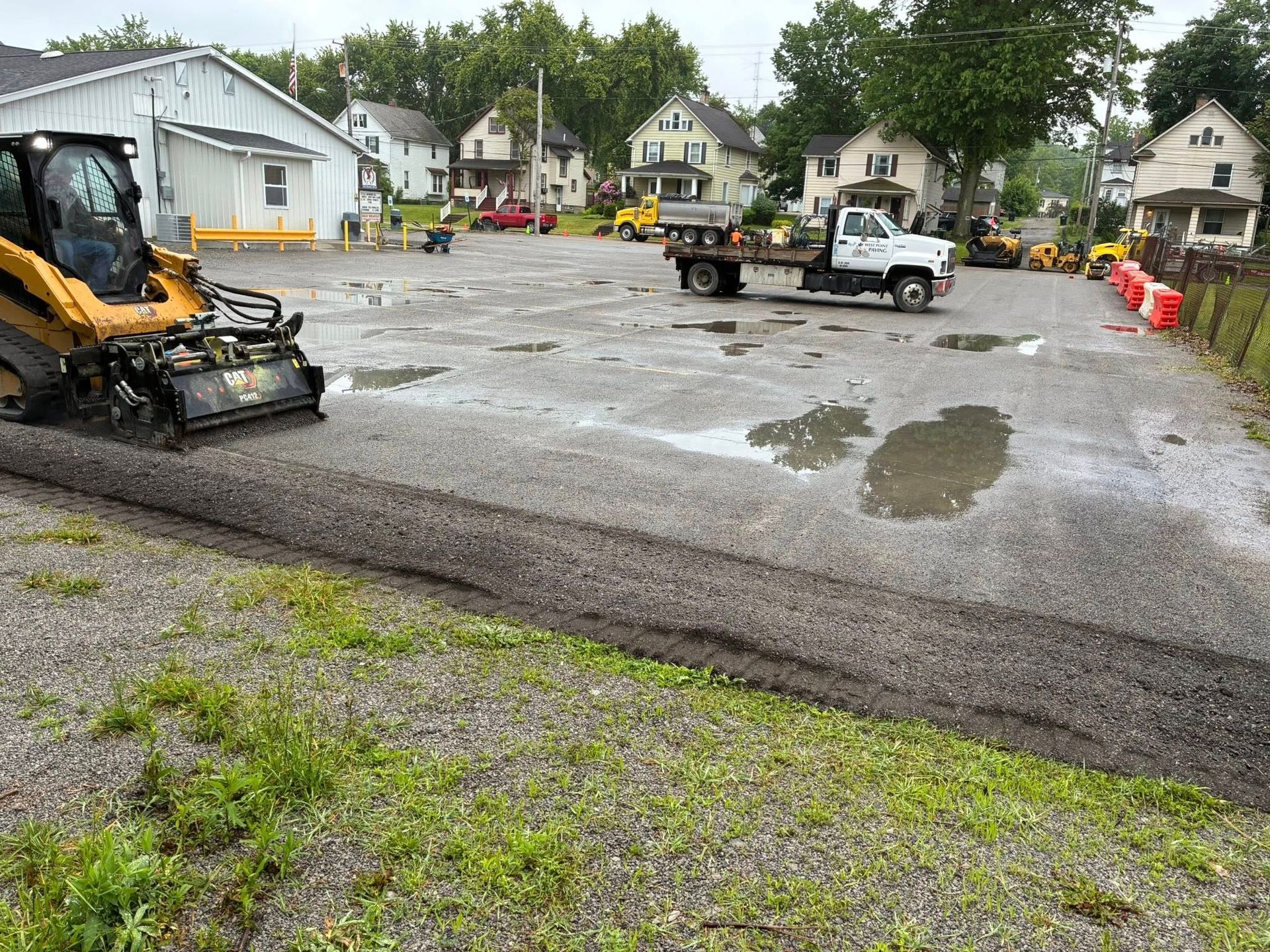 A yellow skid steer removes a section of asphalt from a gravel parking lot, with a pickup truck parked in the background.