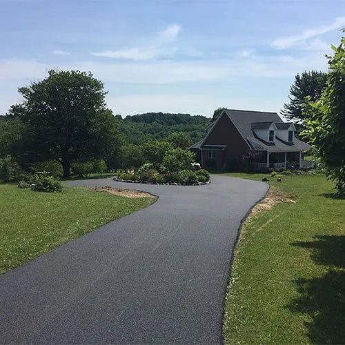 A paved driveway leads toward a house with a dark roof and brick exterior, set against a backdrop of trees and a blue sky.