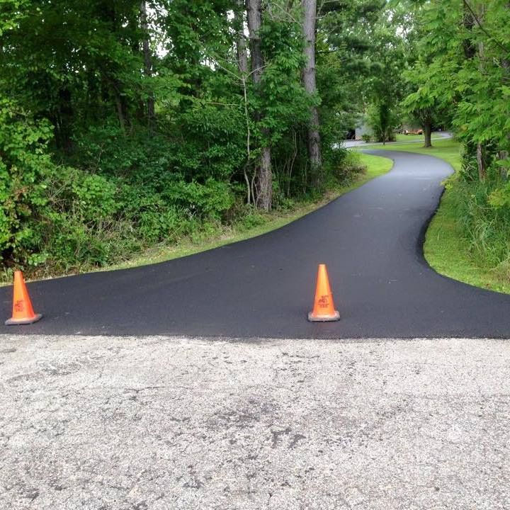 A newly paved, dark asphalt driveway curves into a green, tree-lined area, marked by two orange safety cones at the entrance.