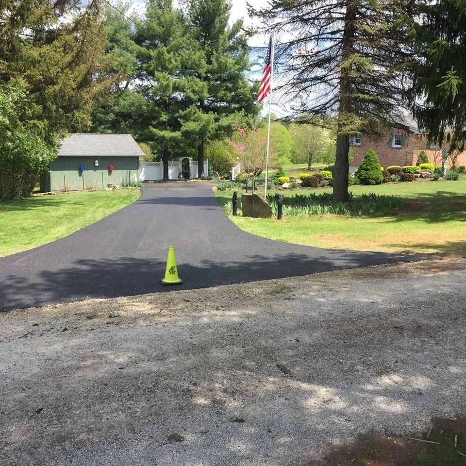 A newly paved black asphalt driveway leads toward a green shed and house, with a yellow traffic cone in the foreground.