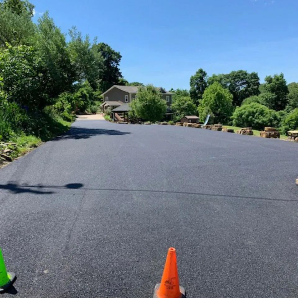 A freshly paved, dark asphalt driveway leads toward a house in the distance, framed by trees and a bright blue sky.
