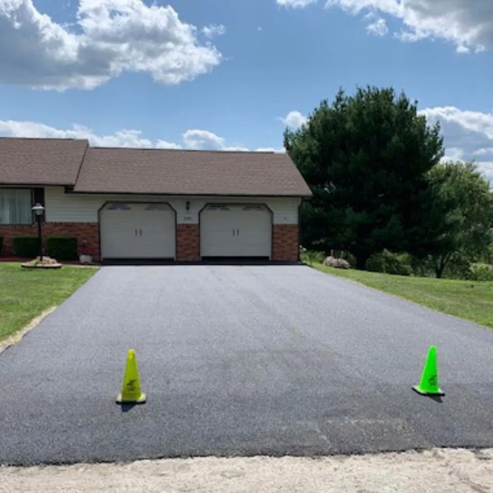 A freshly paved asphalt driveway leading to a two-car garage of a suburban house under a blue, cloudy sky.