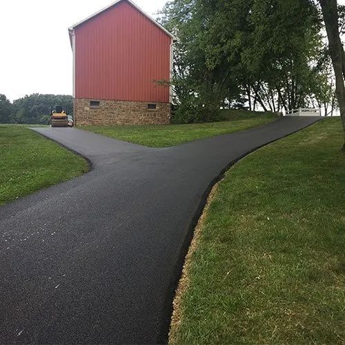 A freshly paved asphalt driveway forks toward a red barn with a stone foundation and a distant roller vehicle.