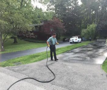 A worker in a light blue shirt uses a hose to apply sealant to a residential driveway.